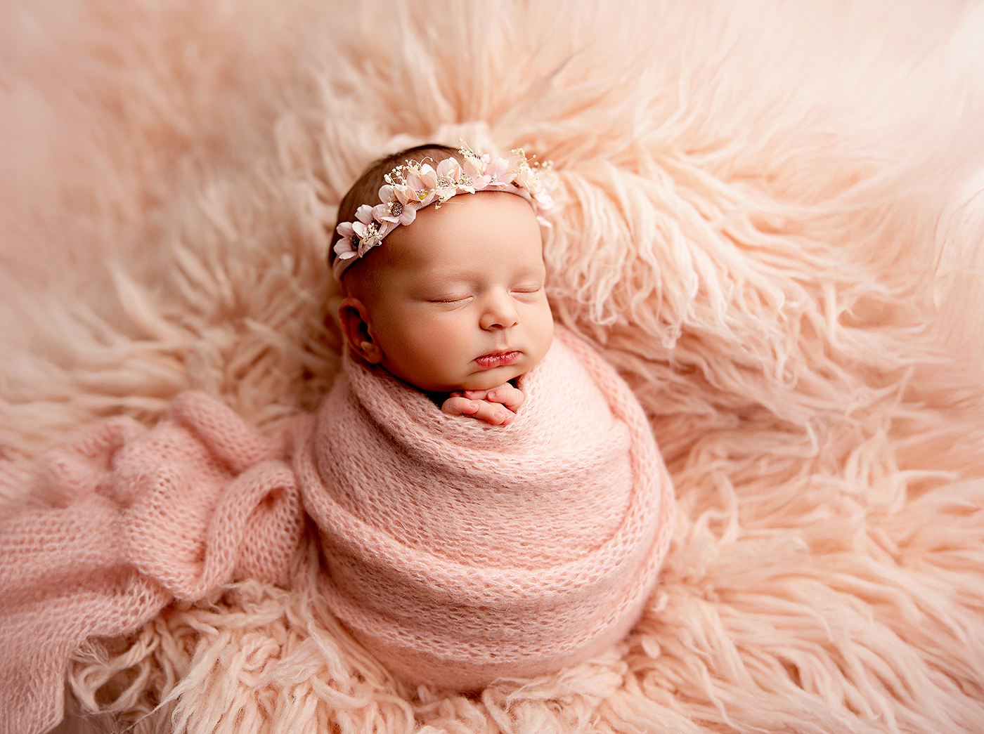 Newborn baby girl swaddled in pink wrap with floral headband, sleeping peacefully on fluffy pink fur backdrop.