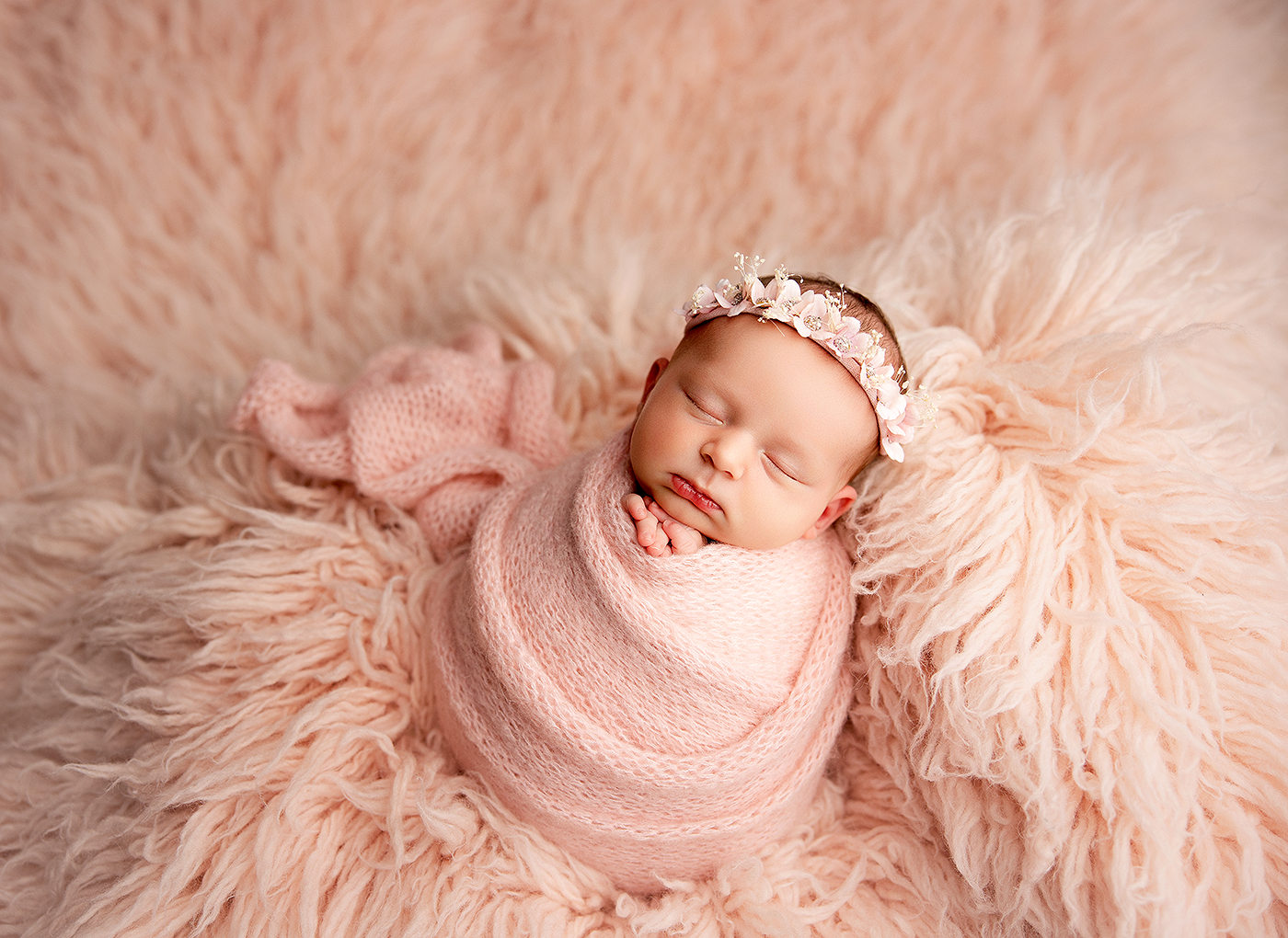Newborn baby girl swaddled in pink wrap sleeping on fluffy blush fur rug wearing floral headband.