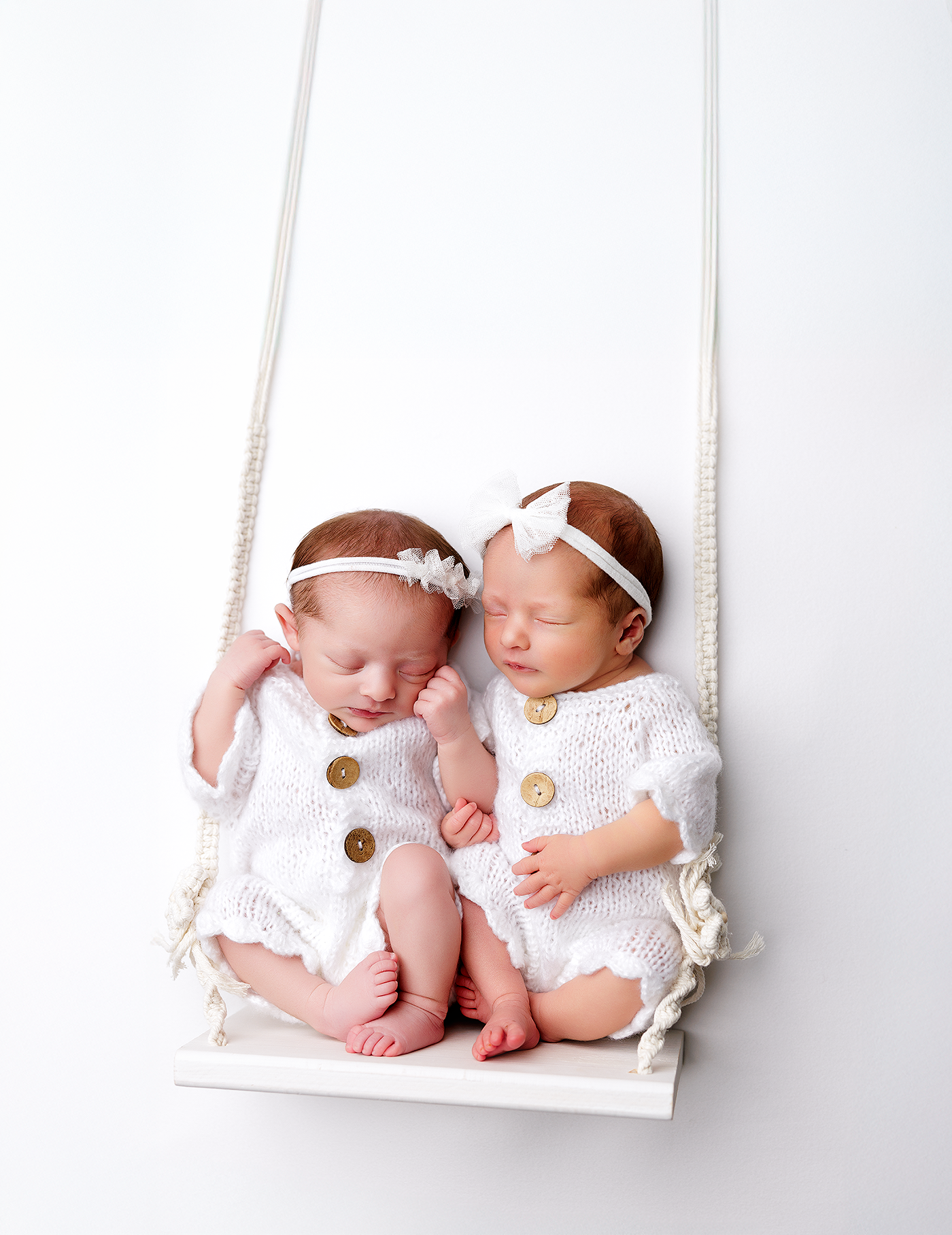 Two sleeping newborn twins in white knit rompers with gold buttons sit together on a small white swing.