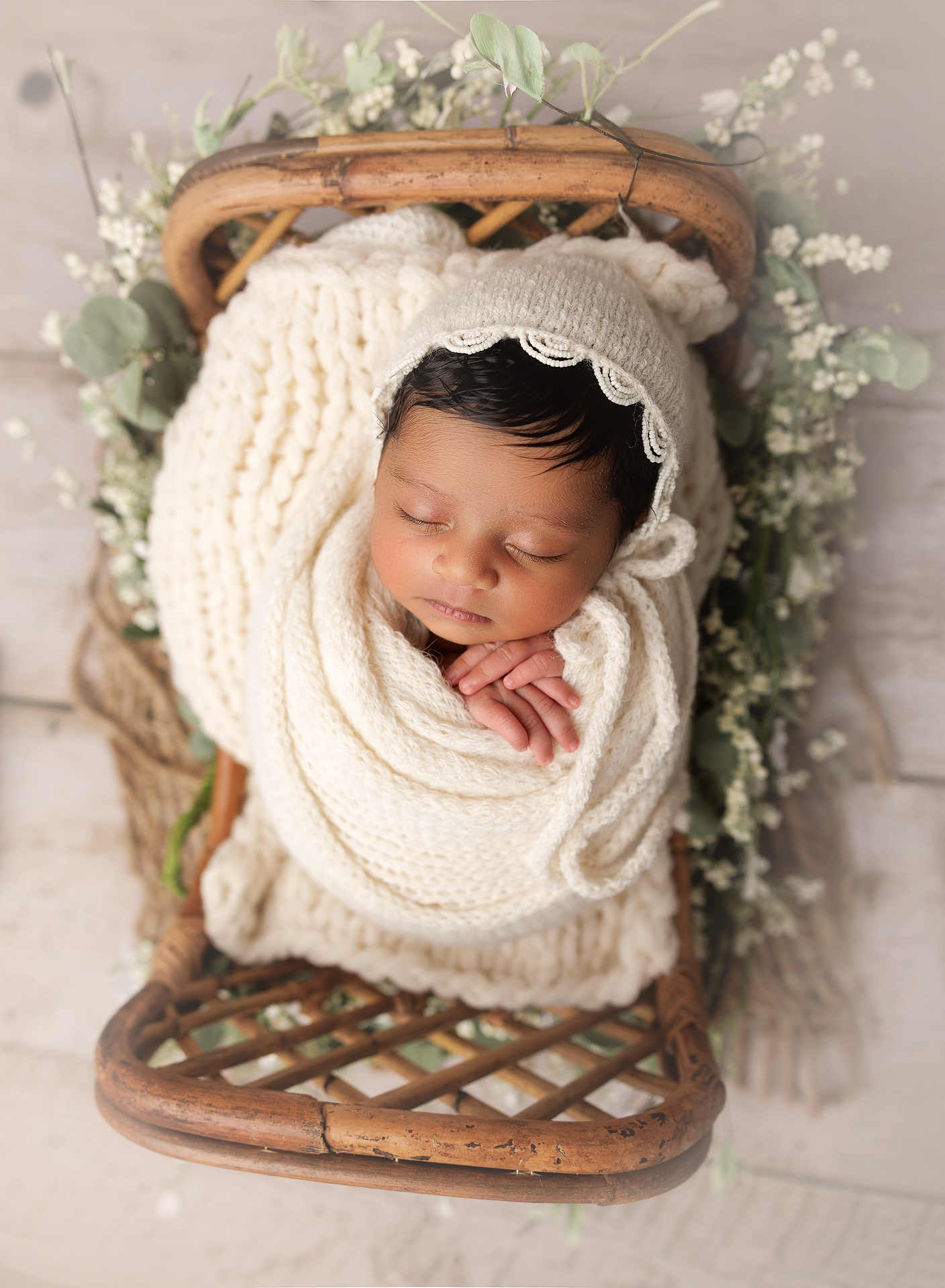 A sleeping newborn wrapped in cream knit blanket nestled in wooden basket with delicate white flowers and greenery.