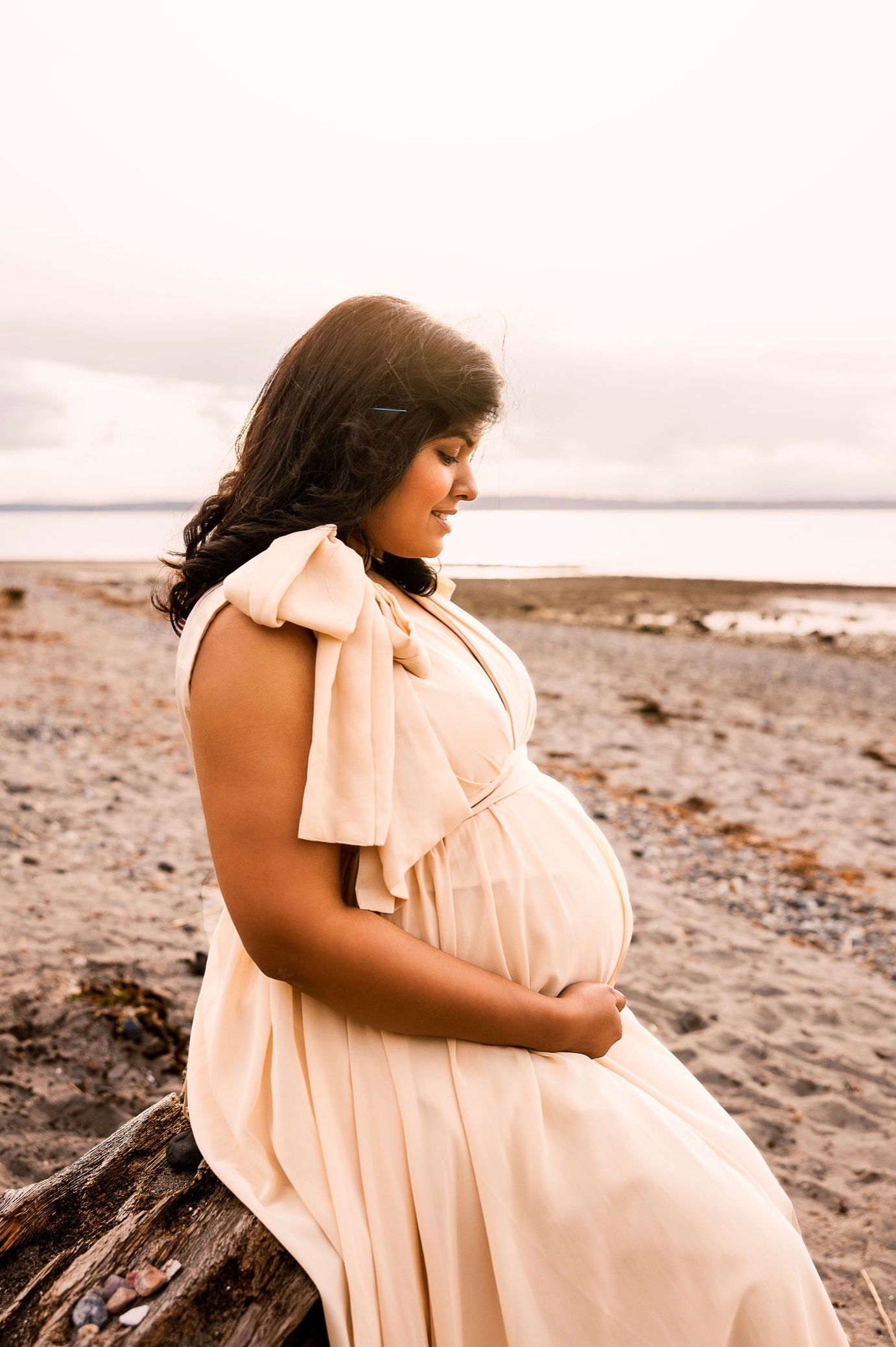 Pregnant woman in cream dress cradling baby bump on rocky beach, maternity photography session at sunset.