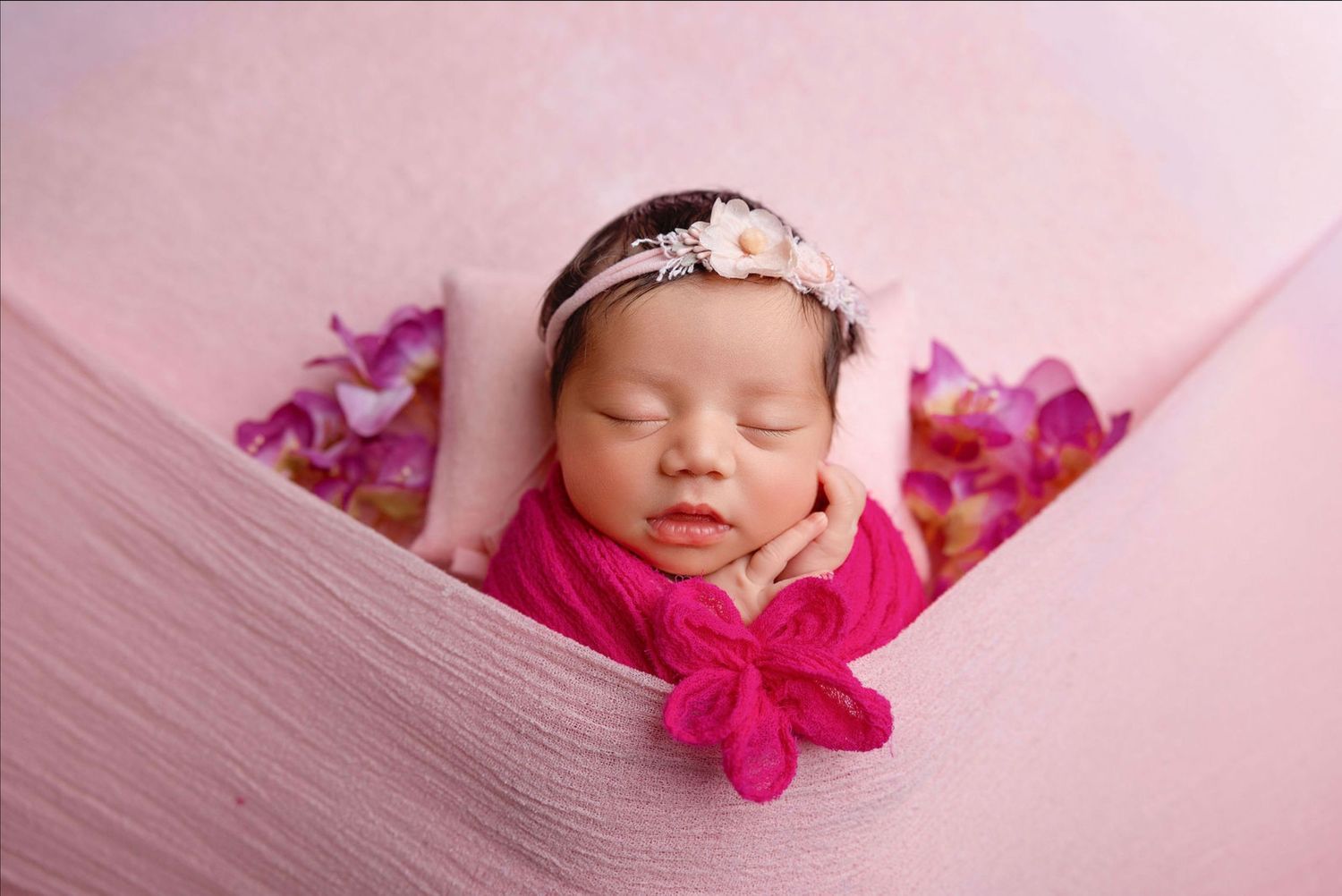 A sleeping newborn baby wearing a pink bow is nestled in soft pink blankets during a professional photo session.
