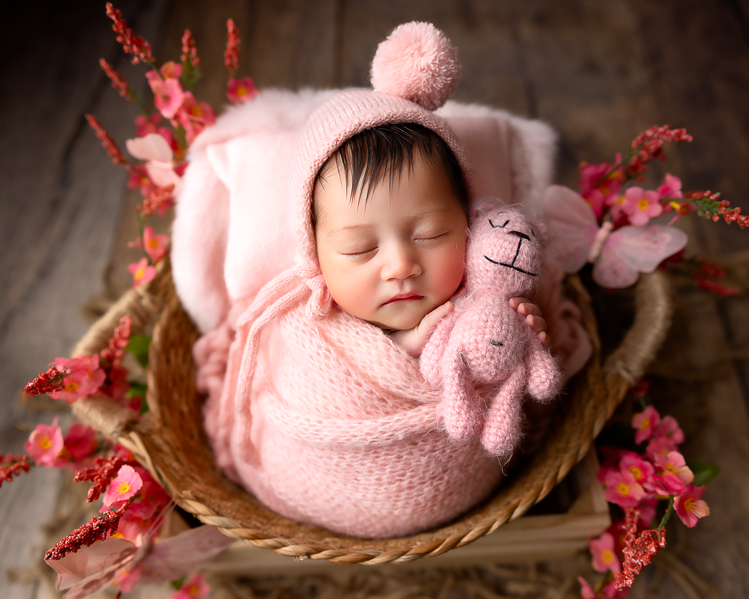 A sleeping newborn in pink swaddle rests peacefully in a rustic basket with red floral accents.
