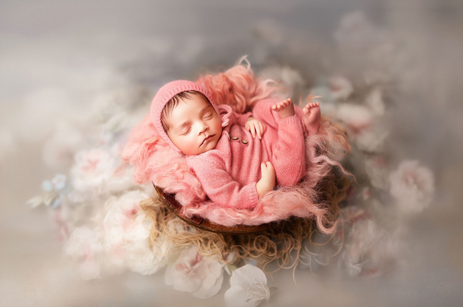 Sleeping newborn baby in pink knit outfit and bonnet nestled in a floral nest for a professional photo.
