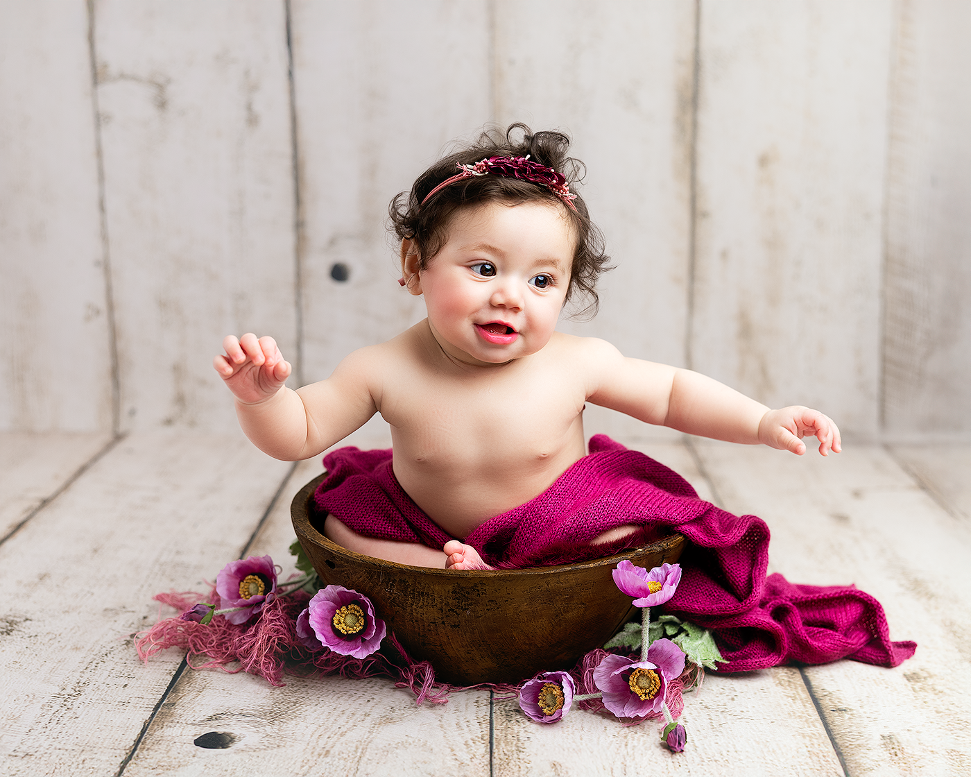 A baby girl sits in a wooden bowl wearing a magenta wrap and floral headband, surrounded by purple flowers on white wood.