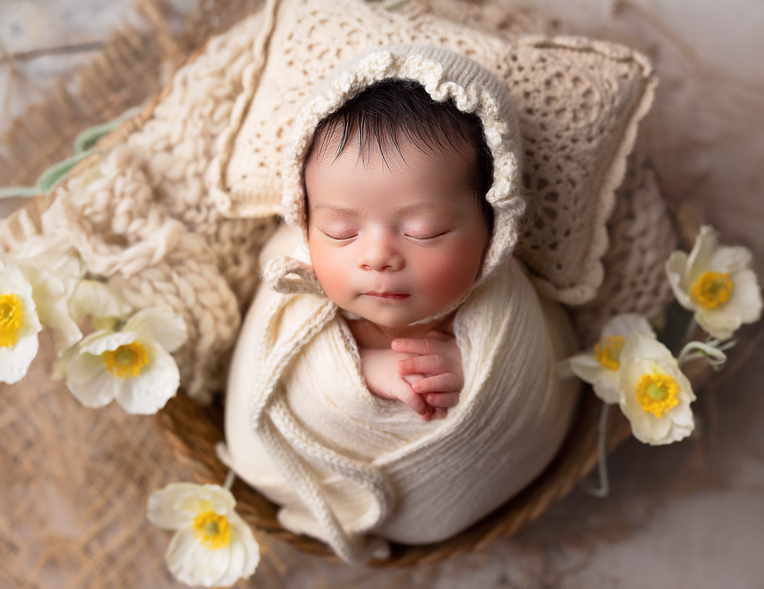 Sleeping newborn wrapped in cream fabric rests on textured pillow with white daffodils surrounding them.