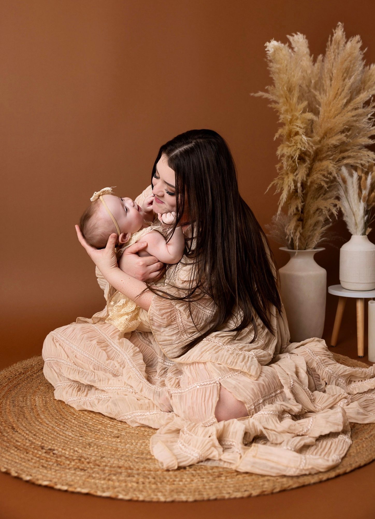 A mother in a beige dress sits on a woven rug, tenderly kissing her baby girl against a warm brown backdrop.