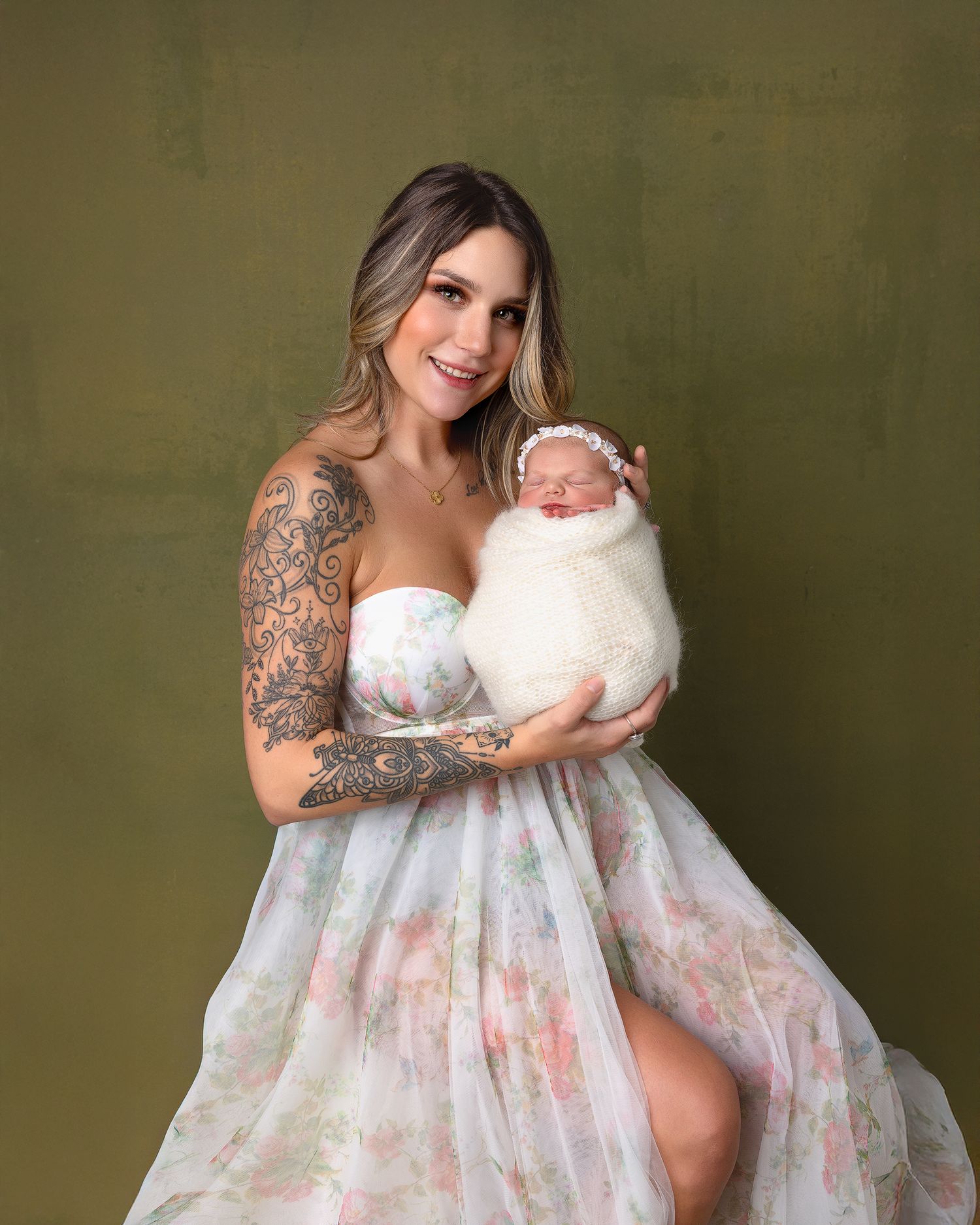 Woman in flowing white and pink floral dress poses against olive green backdrop in maternity photoshoot.