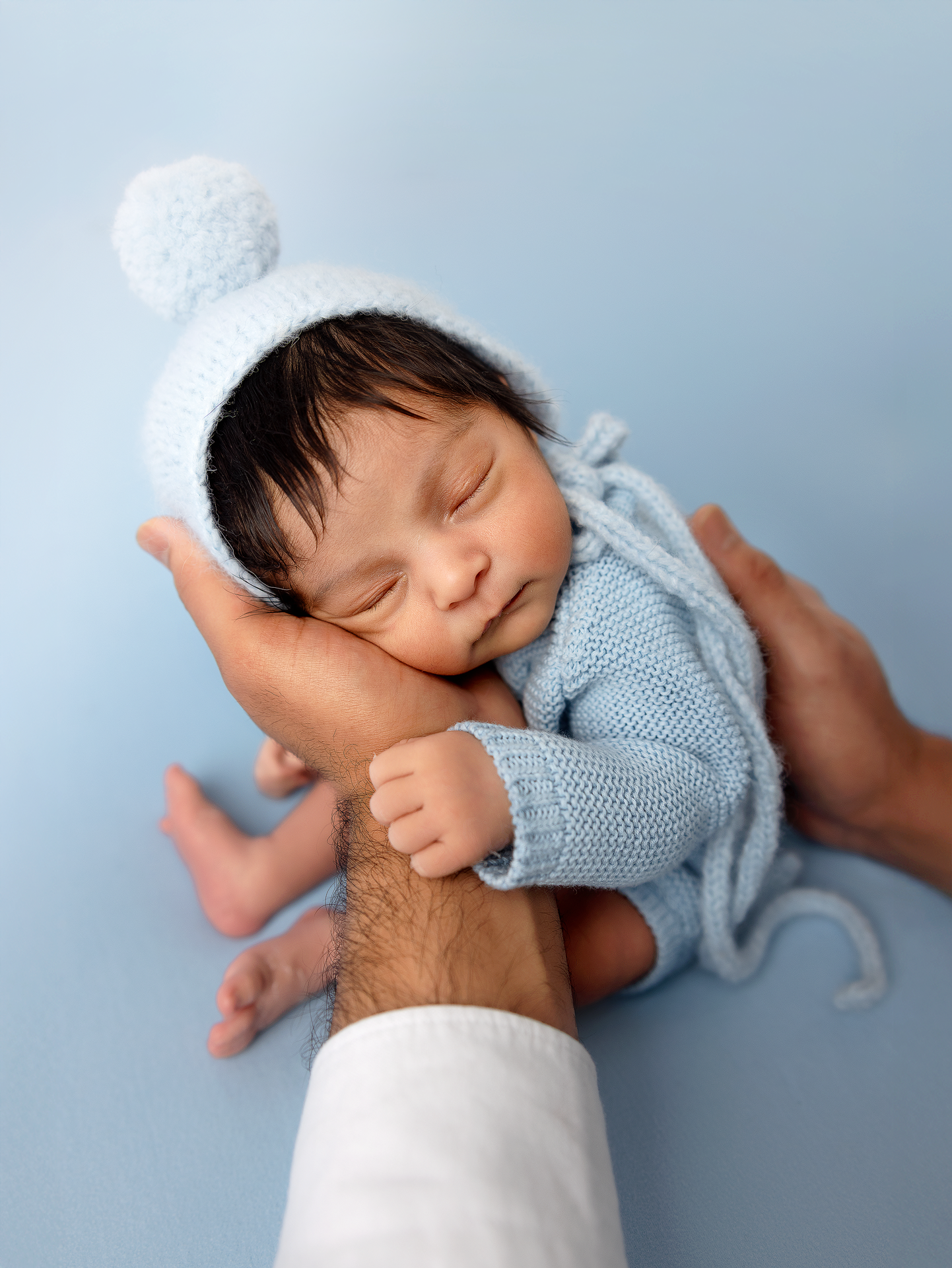 A sleeping newborn baby wearing a blue knit sweater is cradled in gentle hands against a soft blue background.