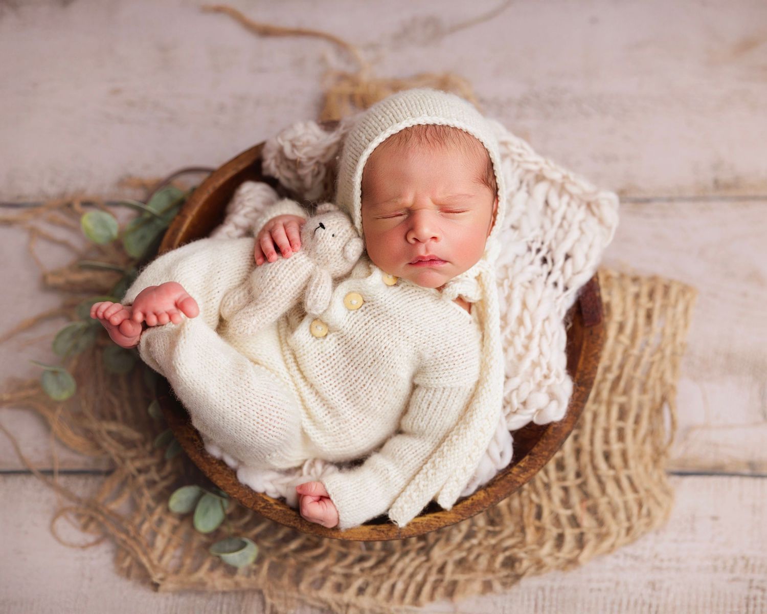 Newborn baby sleeping in a wooden bowl wearing a cream knit outfit and bonnet, holding a white stuffed bunny toy.