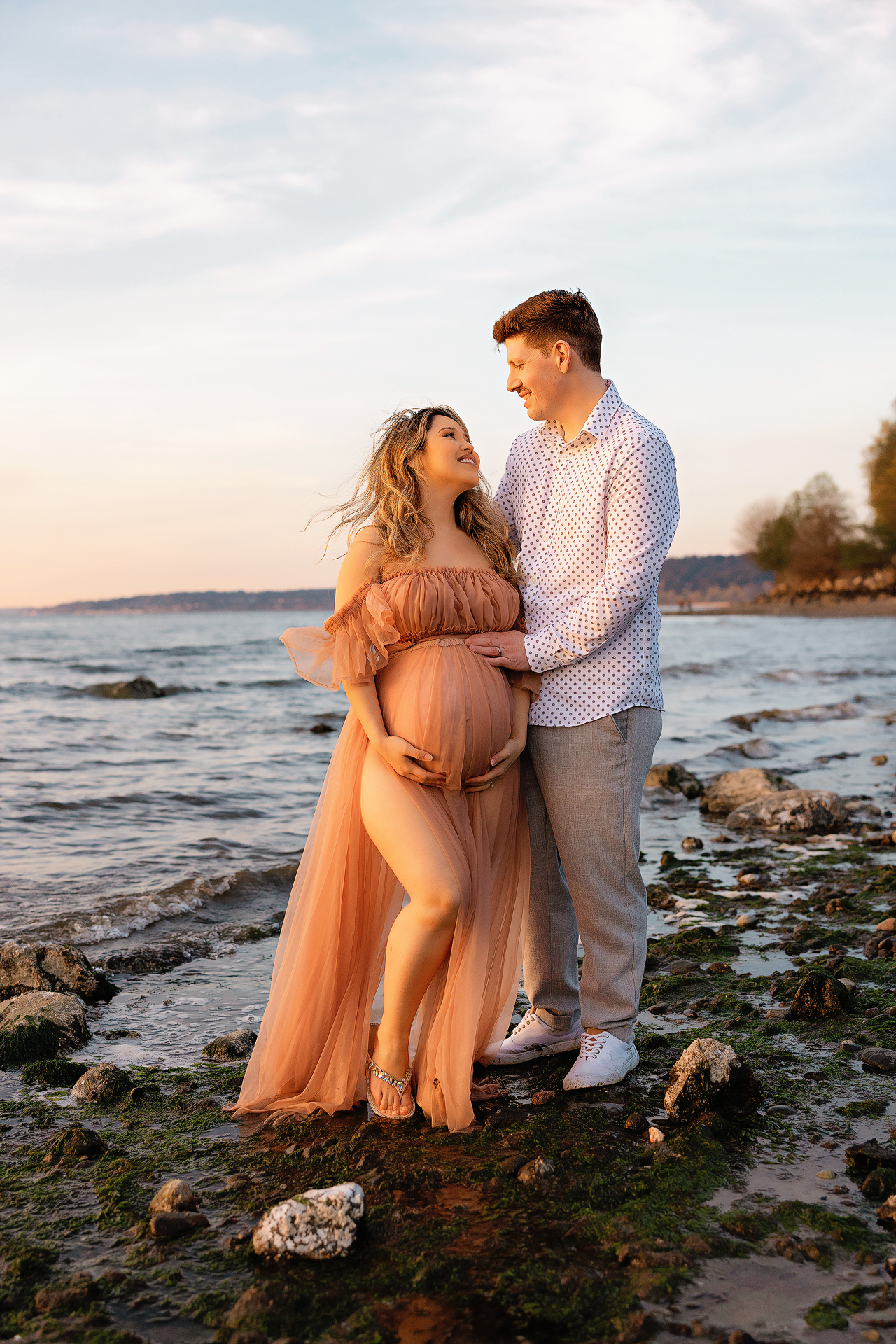 Pregnant couple embracing on rocky shoreline at sunset, woman in flowing peach gown cradling baby bump.