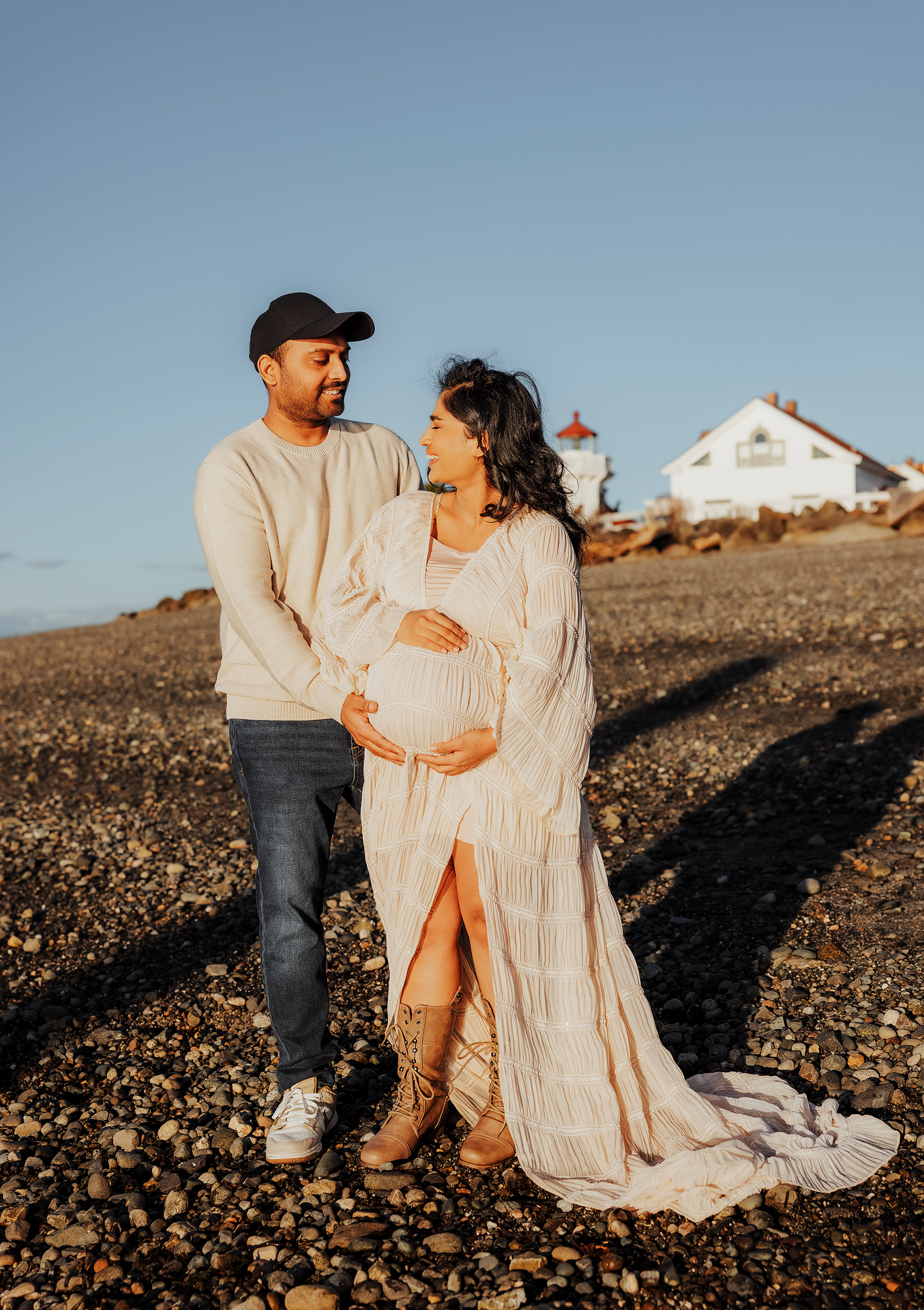Couple poses on rocky beach at golden hour, woman in white maternity gown, man in beige sweater, lighthouse in background.