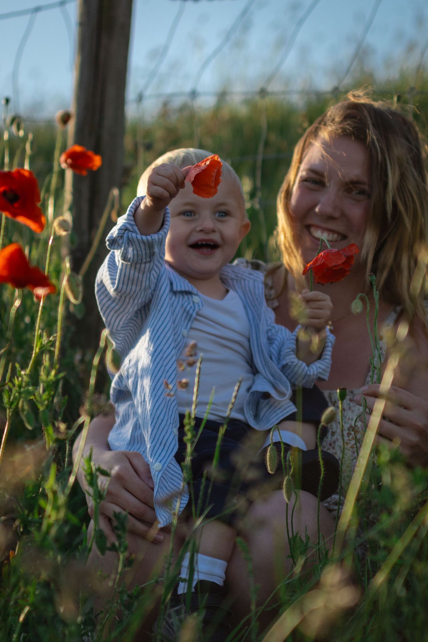 I ett vallmofält sitter en leende mamma och hennes son. Sonen visar glatt upp en vallmoblomma.