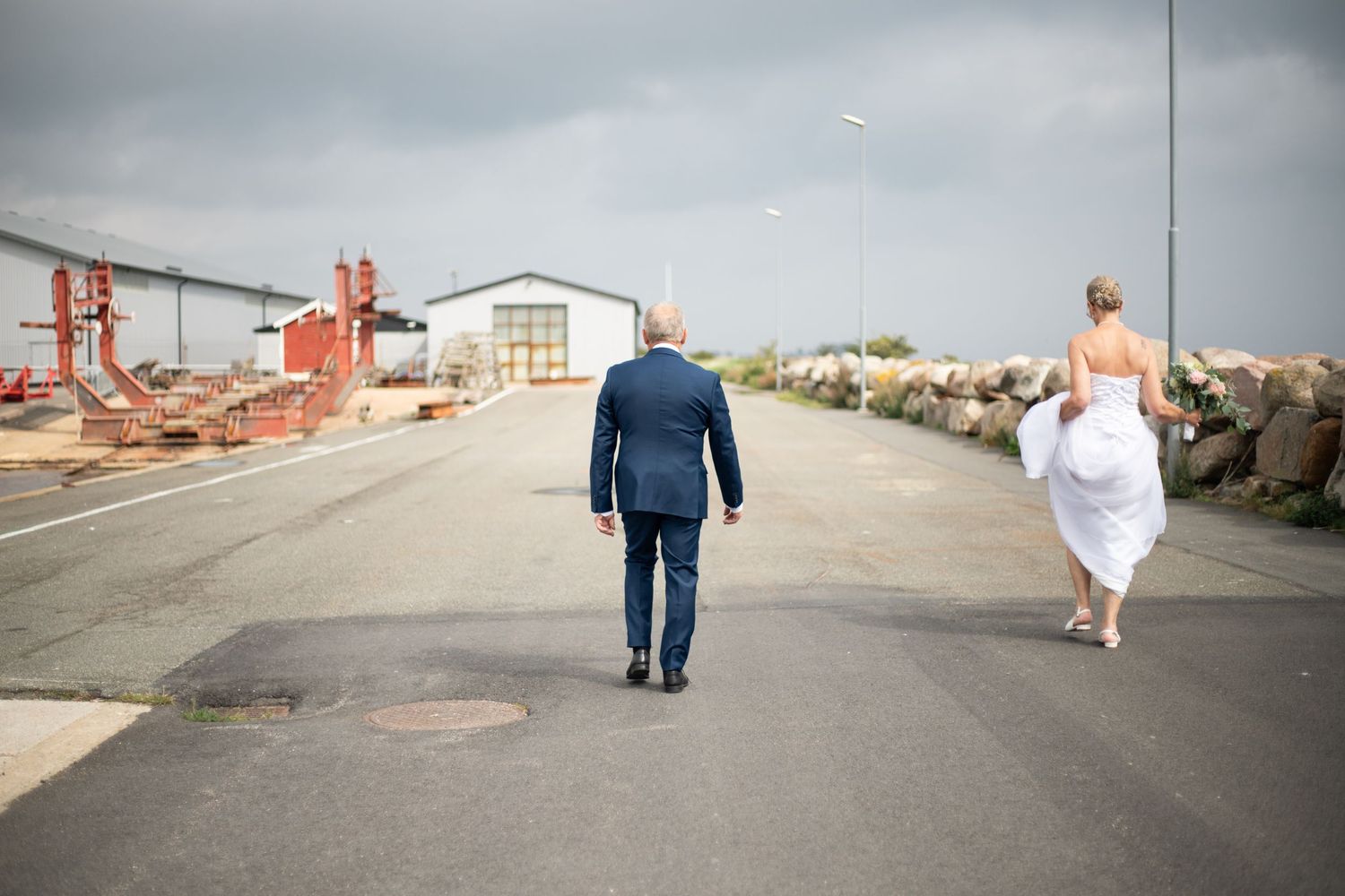 Ett brudpar promenerar bort längs hamnområdet i Skillinge. Kvinnan bär sin klänning i ena handen så att den inte släpar i backen.