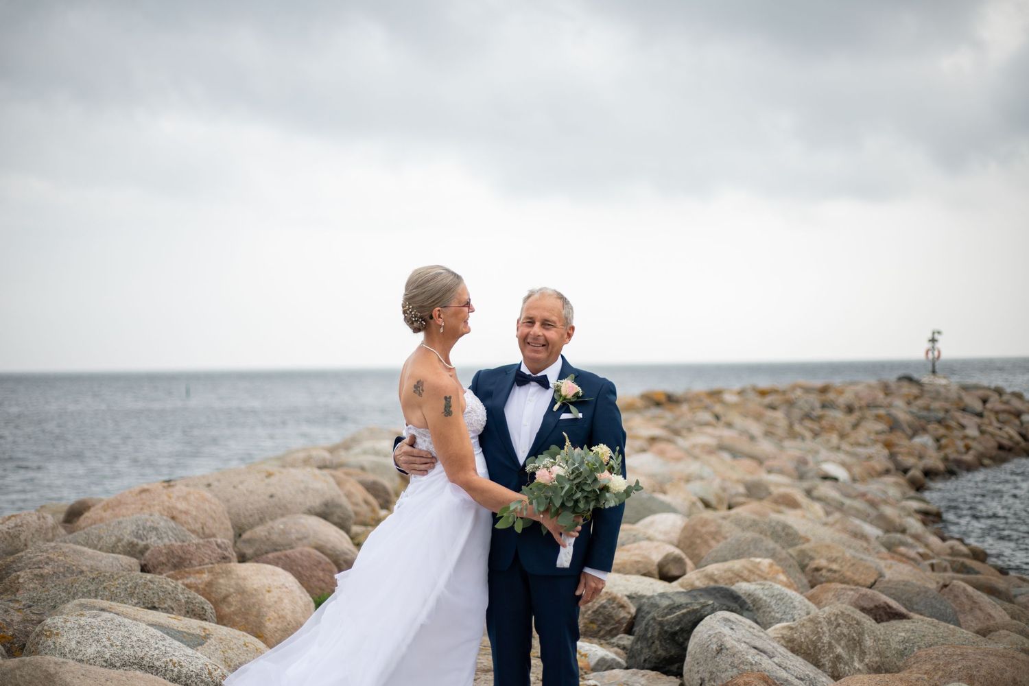 Ett brudpar står vid havet med stenar i bakgrunden. De omfamnar varandra och ser lyckliga ut.