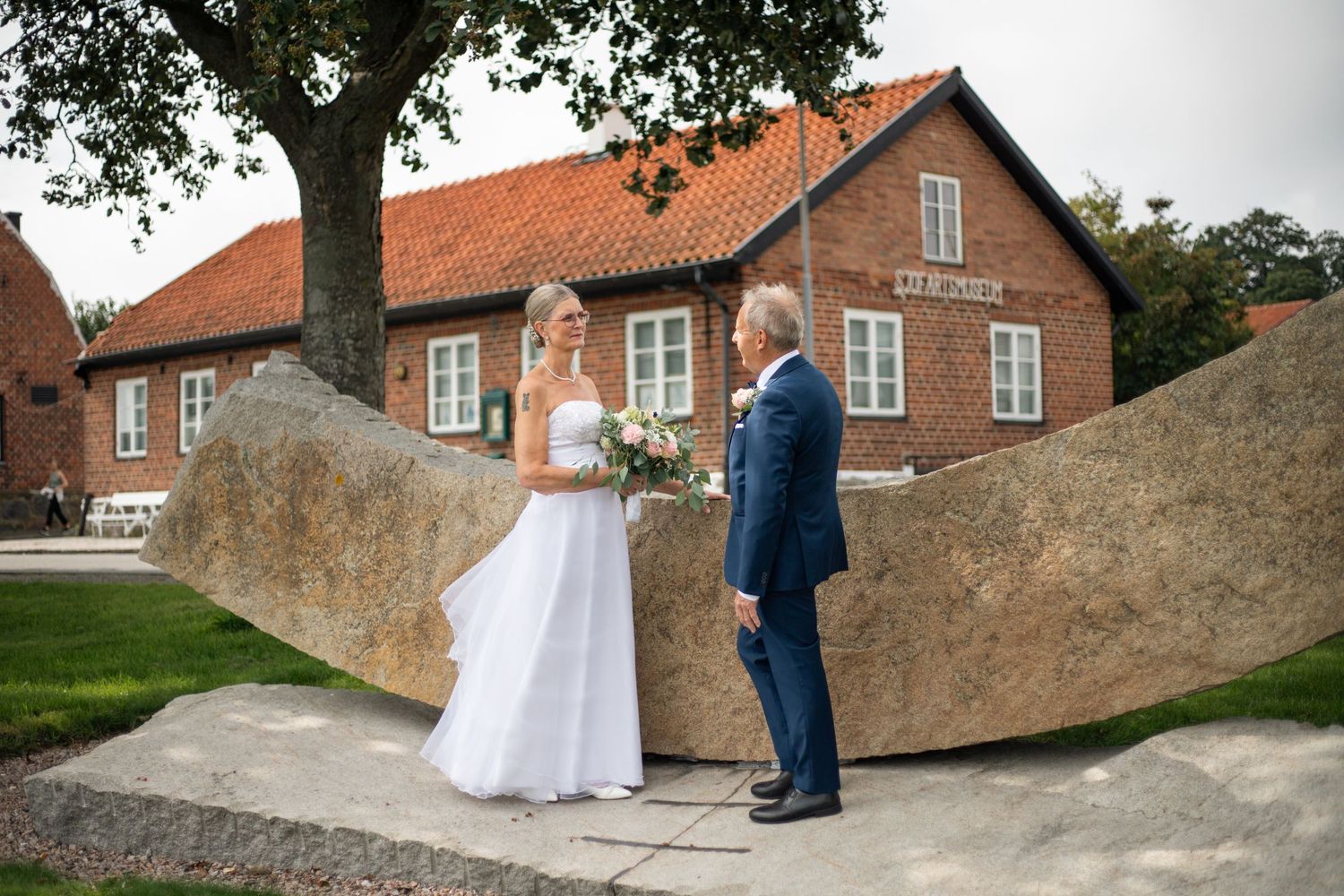 Ett brudpar står vid en staty i Skillinge hus i bakgrunden. Paret tittar kärleksfullt på varandra.