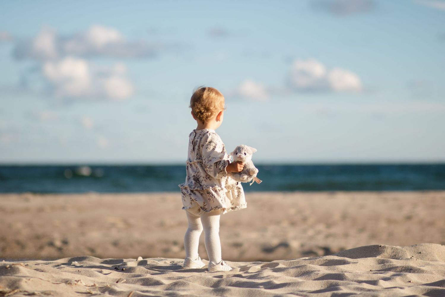 På en strand med havet i bakgrunden står en liten flicka. Hon iklädd ljus klänning och ljusa strumpbyxor. I handen håller hon ett gosedjur.