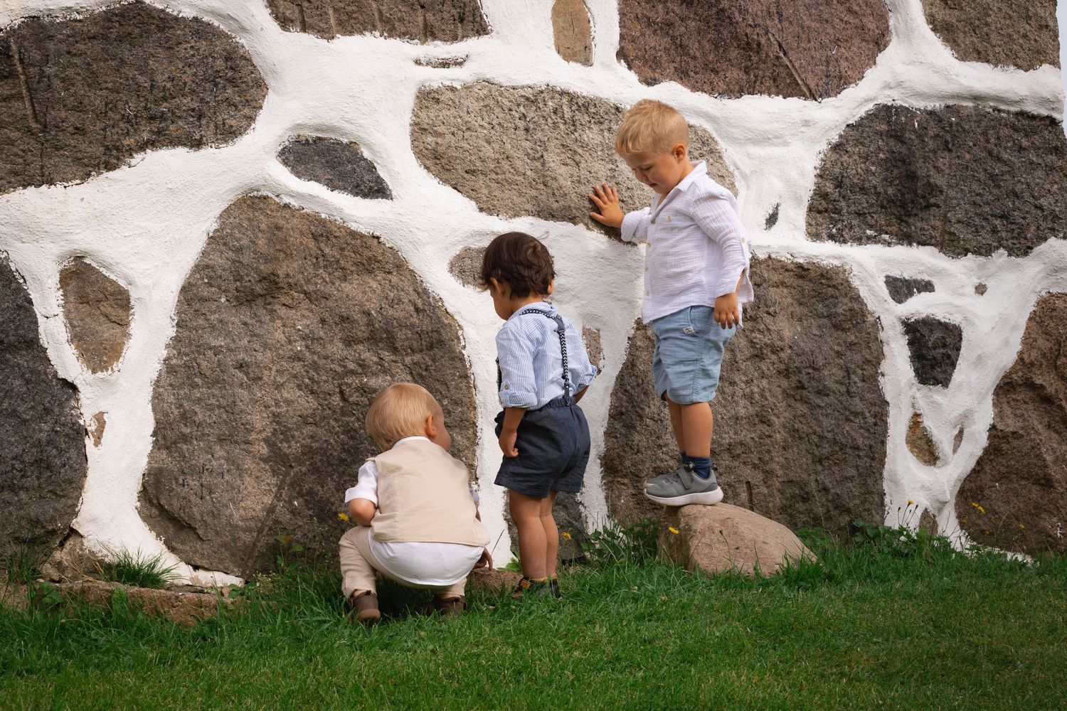 Tre små barn står i tripp trapp trull mot en stenvägg. Den minsta sitter på huk och det tredje barnet står på en sten.