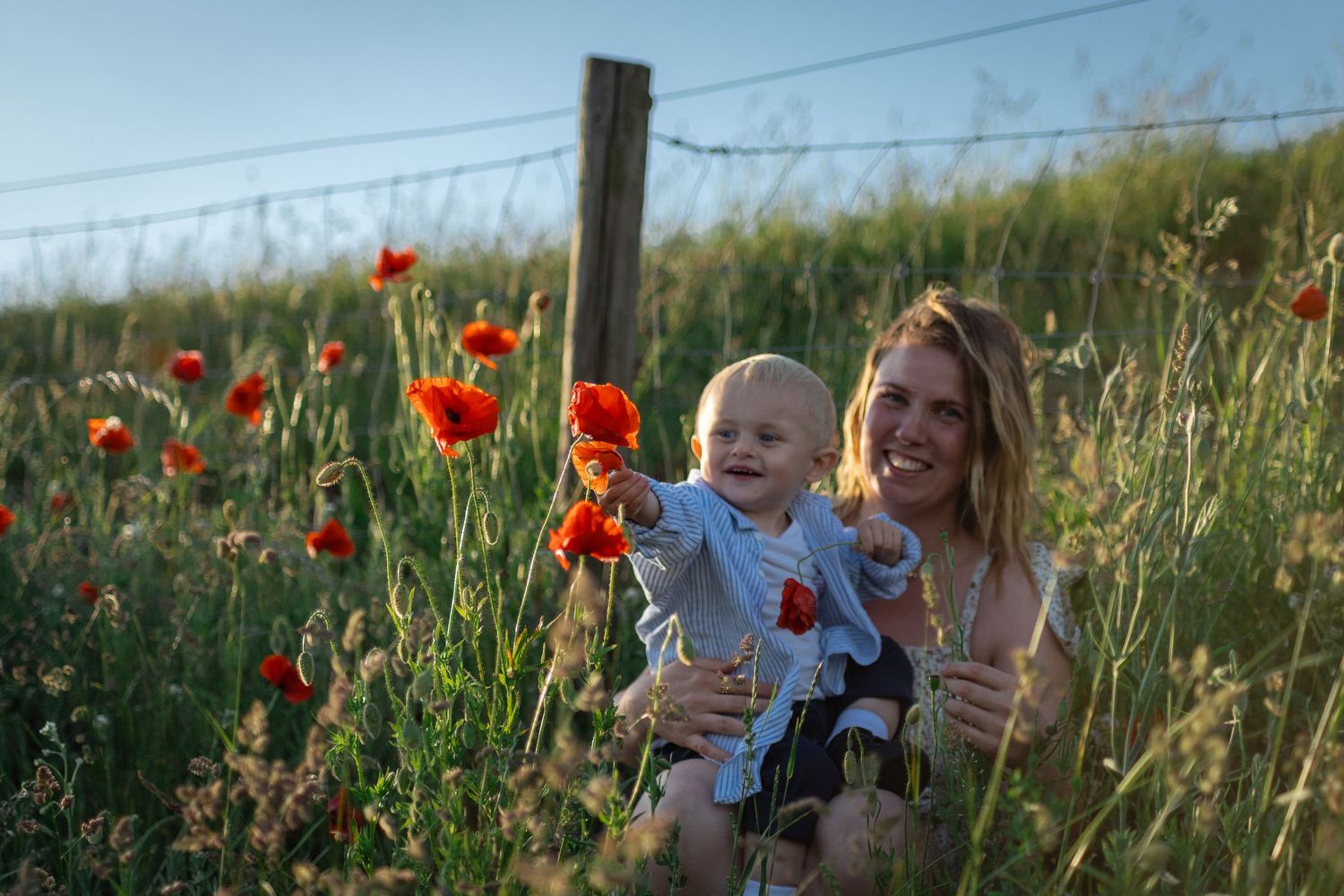 I ett vallmofält sitter en leende mamma och hennes son. Sonen sträcker sig mot vallmoblommorna.