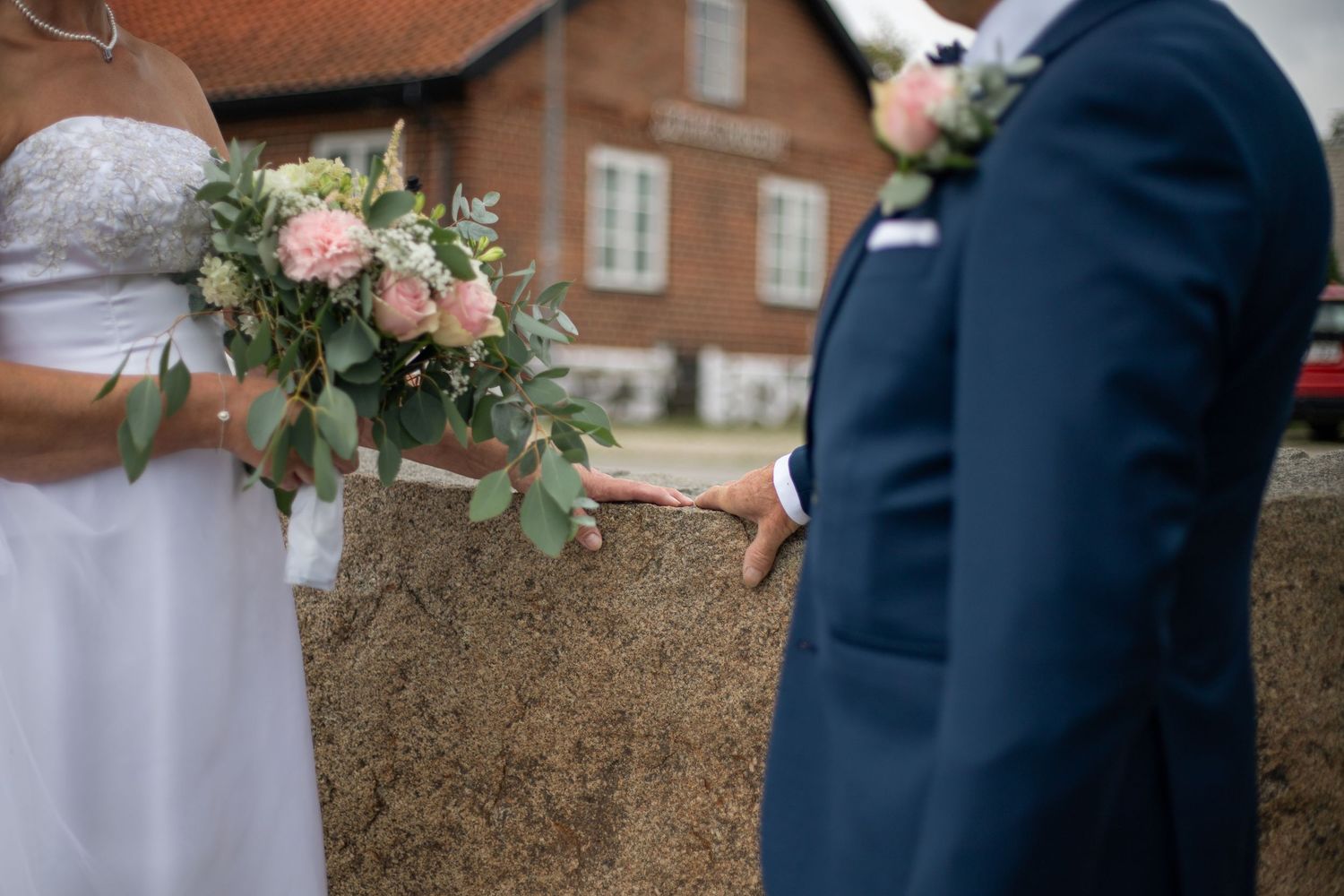 En inzoomad bild på ett brudpar där fokus är på deras händer som vilar tillsammans på en sten. Kvinnan håller i sin brudbukett.