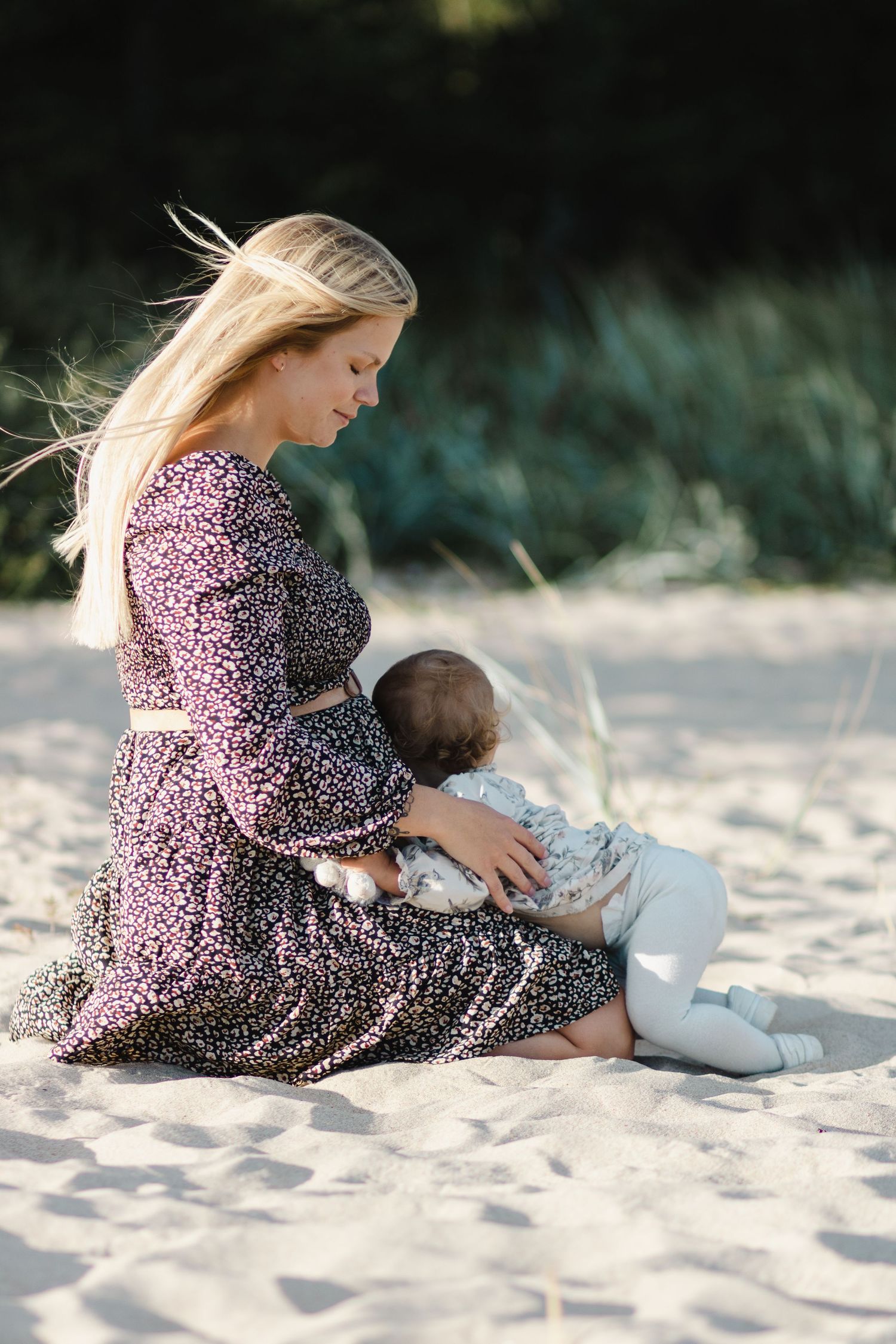 På en strand sitter en mamma. I famnen lutar sig hennes lilla dotter mot den gravida magen.