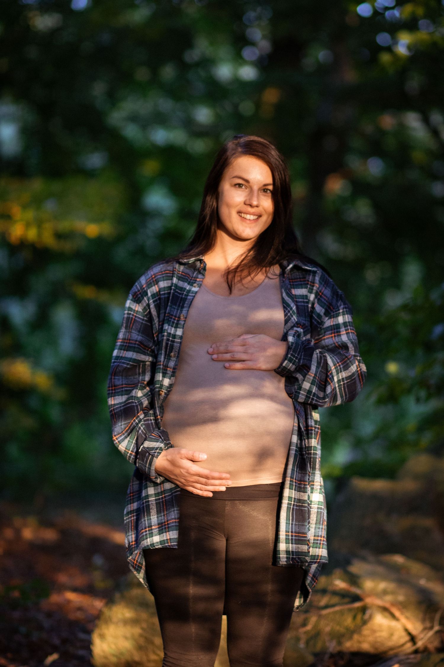 En gravid mamma står och kollar mot kameran i kvällsljuset.