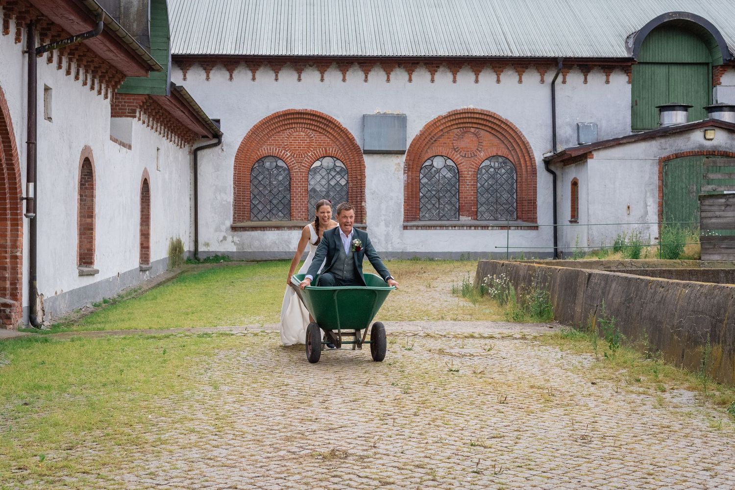 På en innergård kommer en brud körande på brudgummen som sitter i en skottkärra.