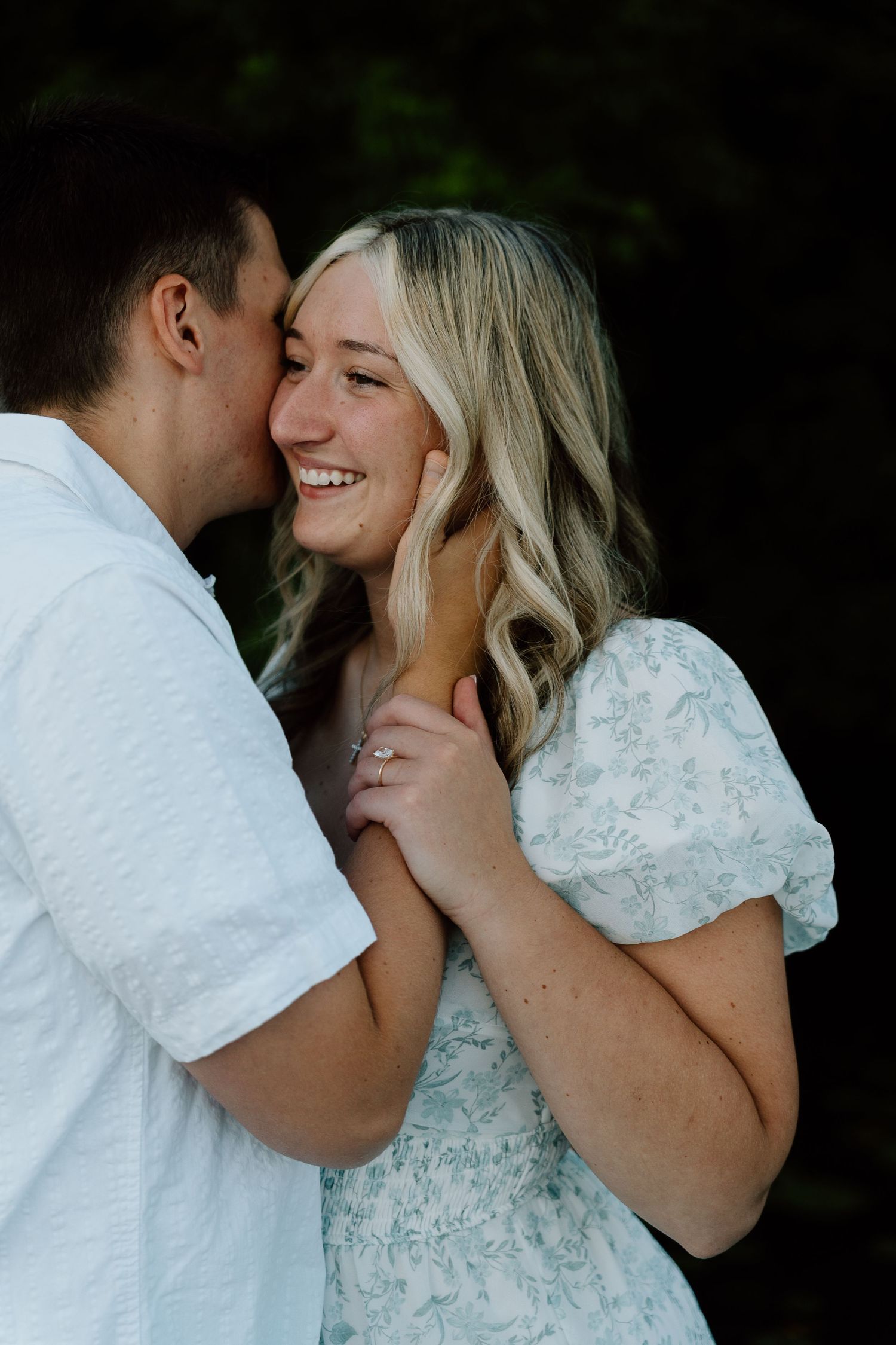 A smiling blonde woman in a floral dress receives a kiss on the cheek from a man in a white shirt outdoors.