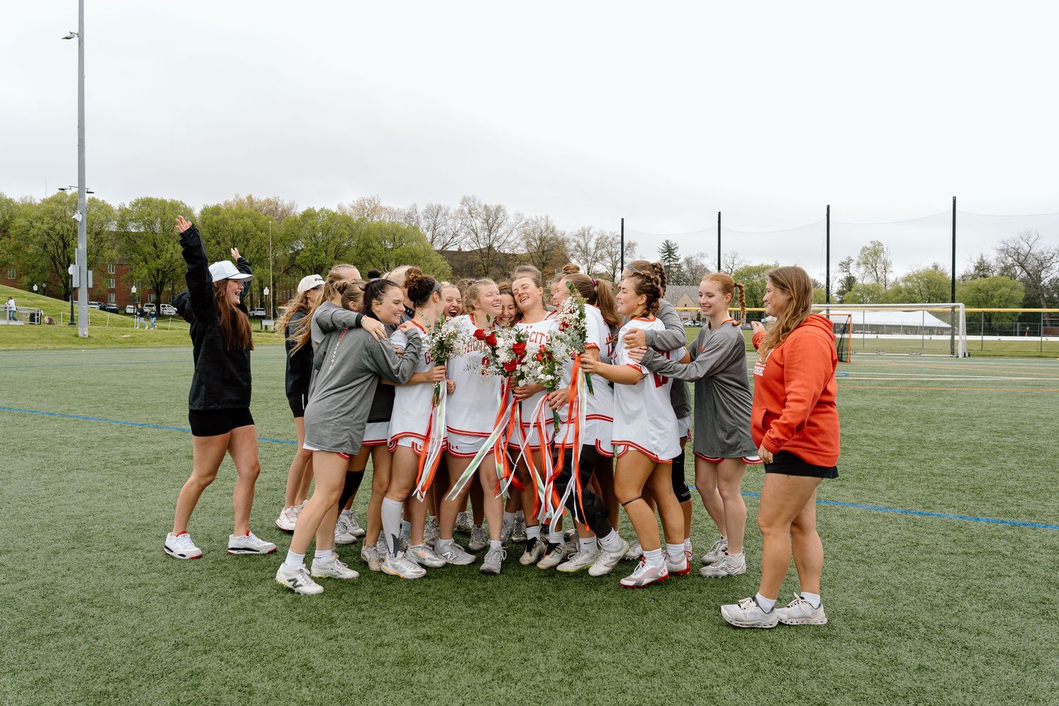 Girls lacrosse team celebrating on green field, huddled together with ribbons and flowers after a victory.