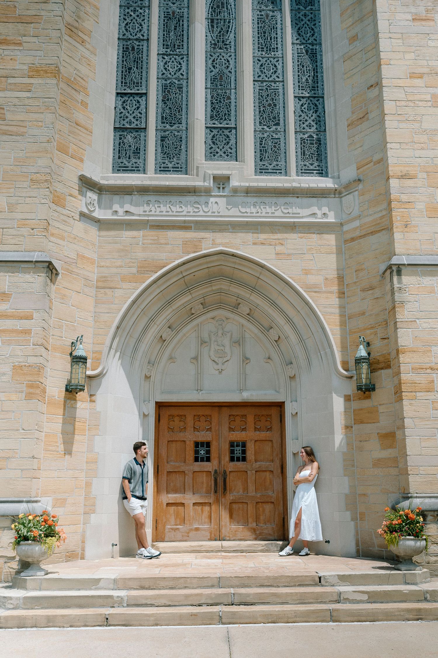 Ornate church entrance with stone walls, stained glass window, and wooden doors flanked by standing couple on the steps.