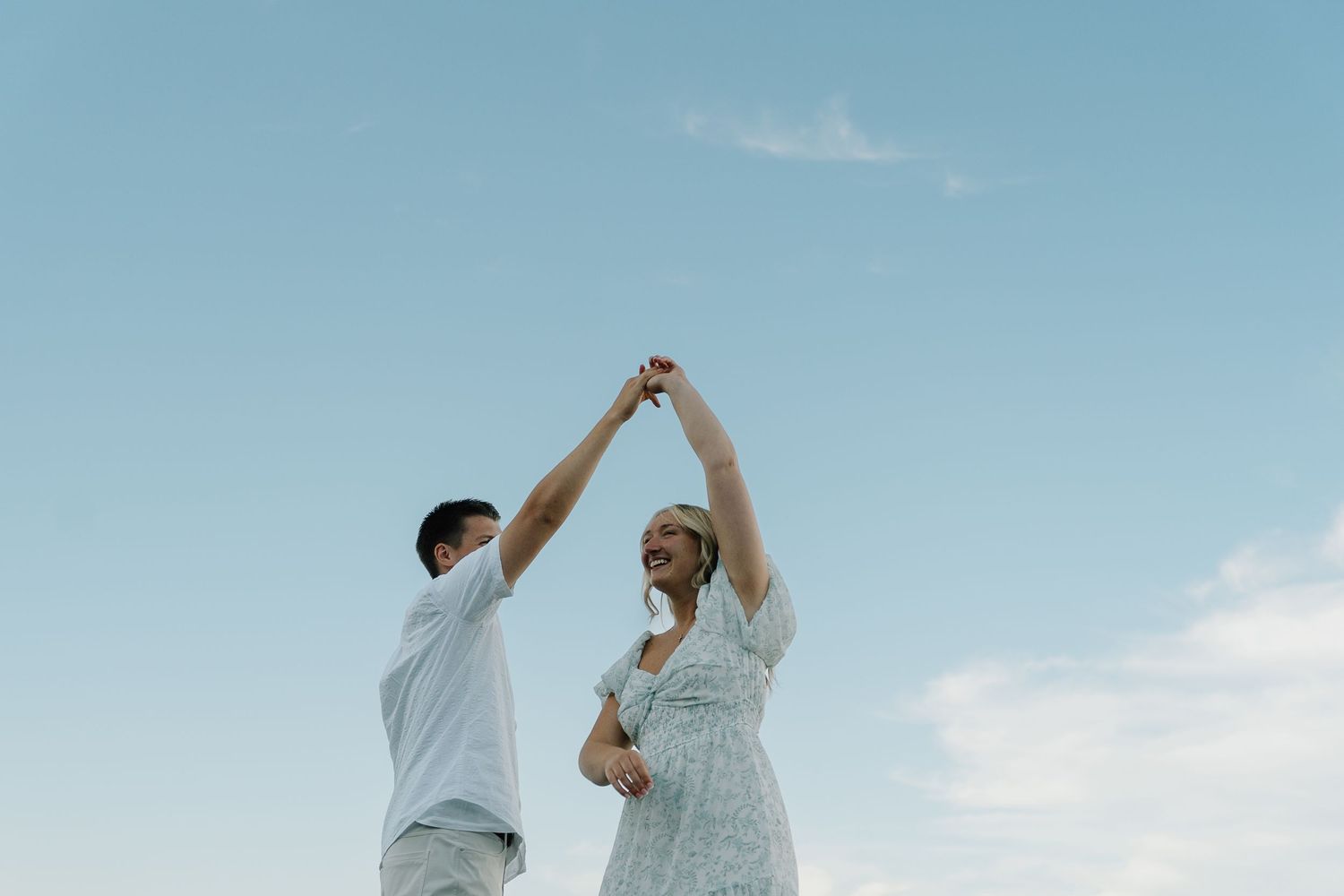 Couple dancing joyfully under a blue sky, raising their hands together in a romantic outdoor moment.