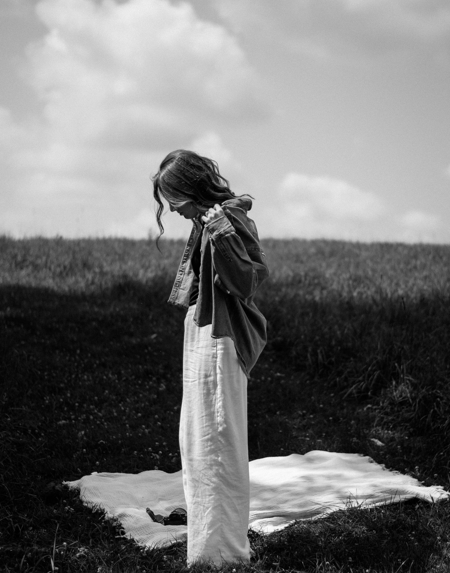 A figure in a white dress stands in a field against a moody sky in black and white.
