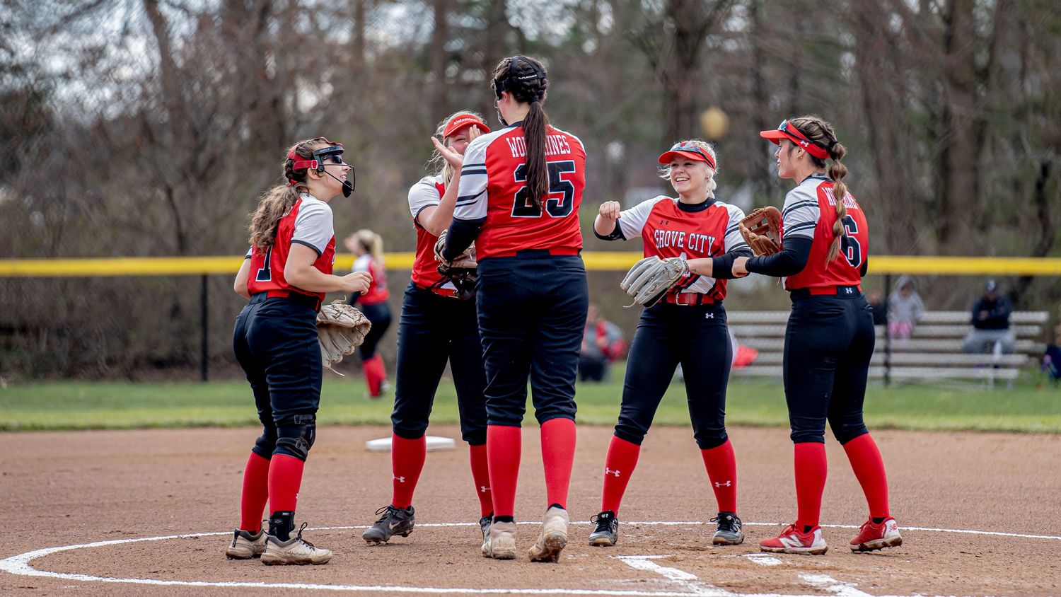 Softball players in red and black uniforms gathered for team discussion on baseball diamond.