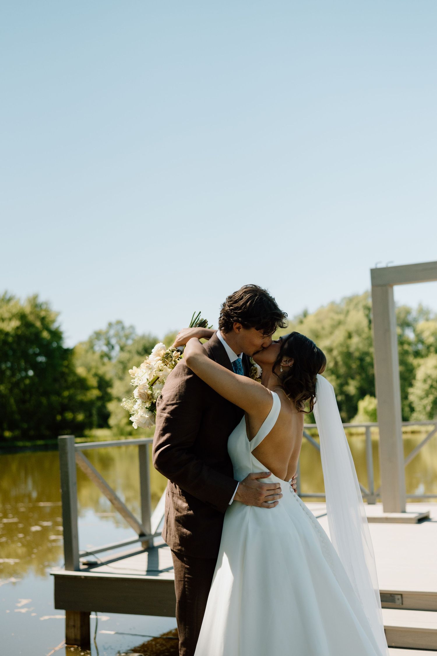 Bride and groom share a romantic kiss on a lakeside dock, bride holding white bouquet in a backless gown on sunny day.