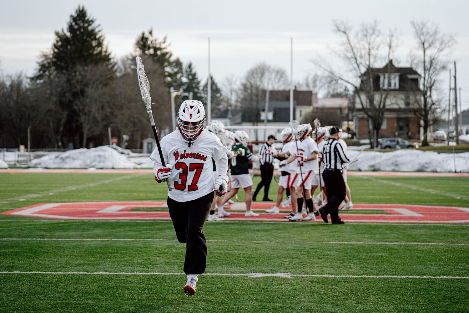 Lacrosse players compete on a snowy field during a winter match as spectators watch from the sidelines.