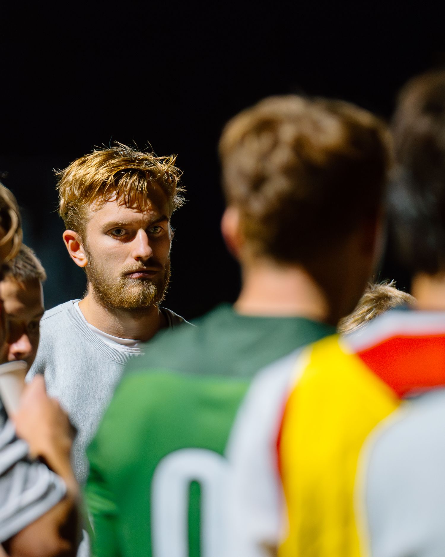 Grove City Men's Soccer team members engaged in a huddle during a nighttime game, with players in colored jerseys.
