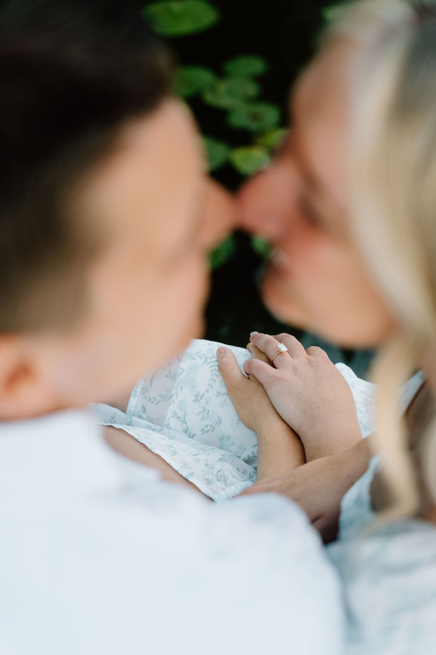 Couple sharing a tender nose-to-nose moment while holding hands, woman wearing an engagement ring, soft focus romantic photo.
