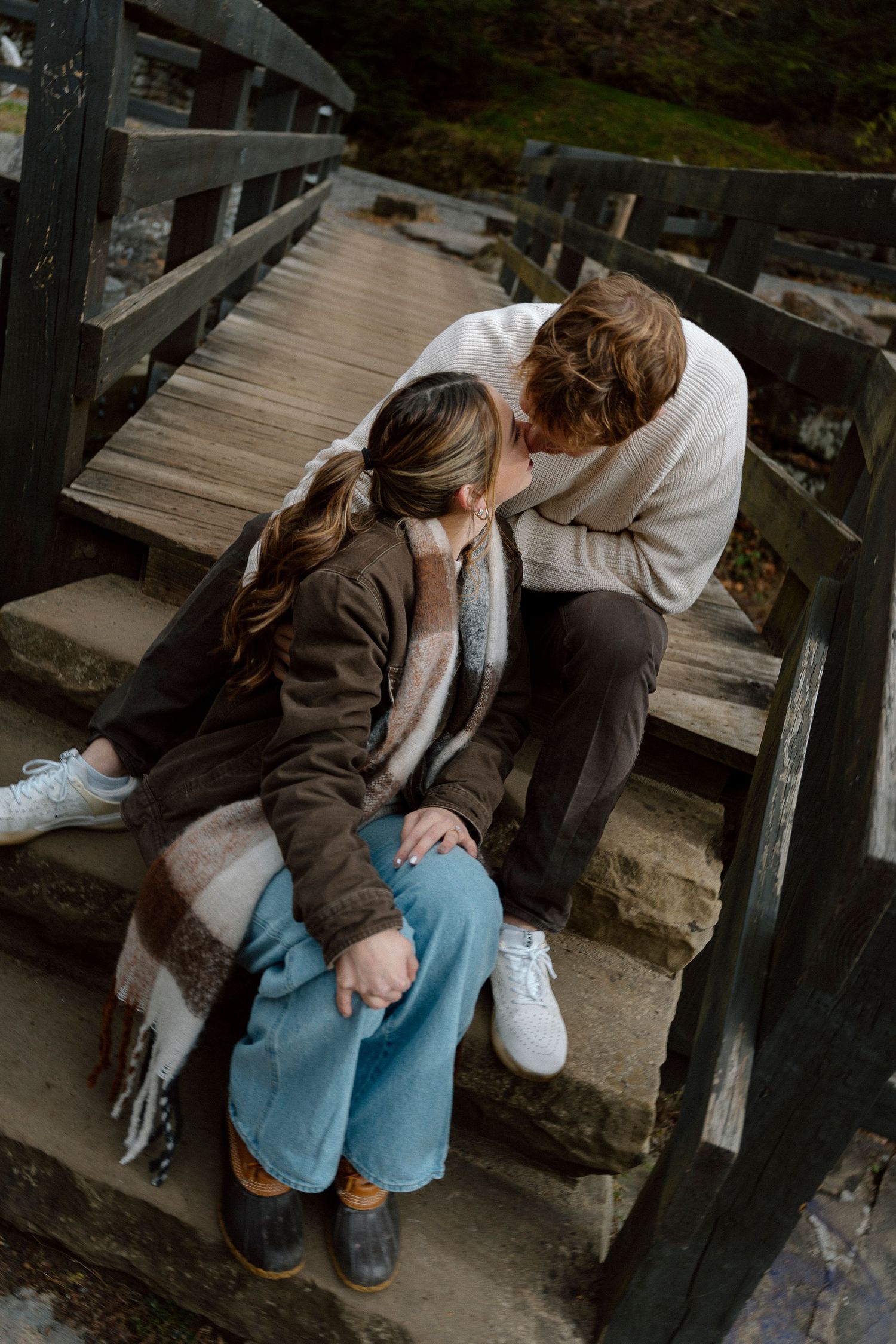Person in brown jacket and jeans sits contemplatively on wooden boardwalk steps in natural setting.