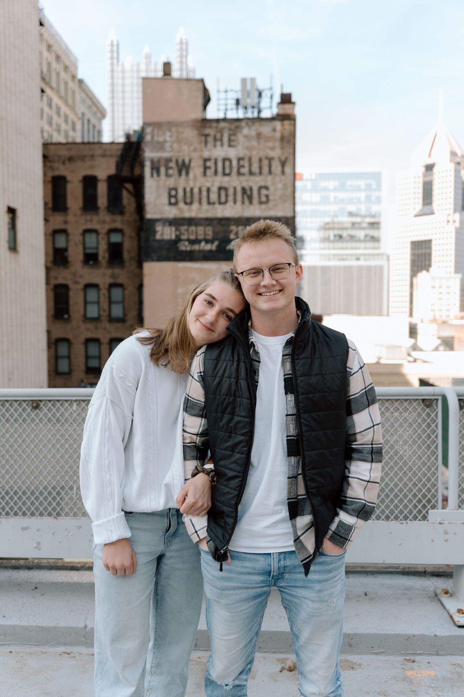 A casual couple posing together on a rooftop with urban architecture in the background.