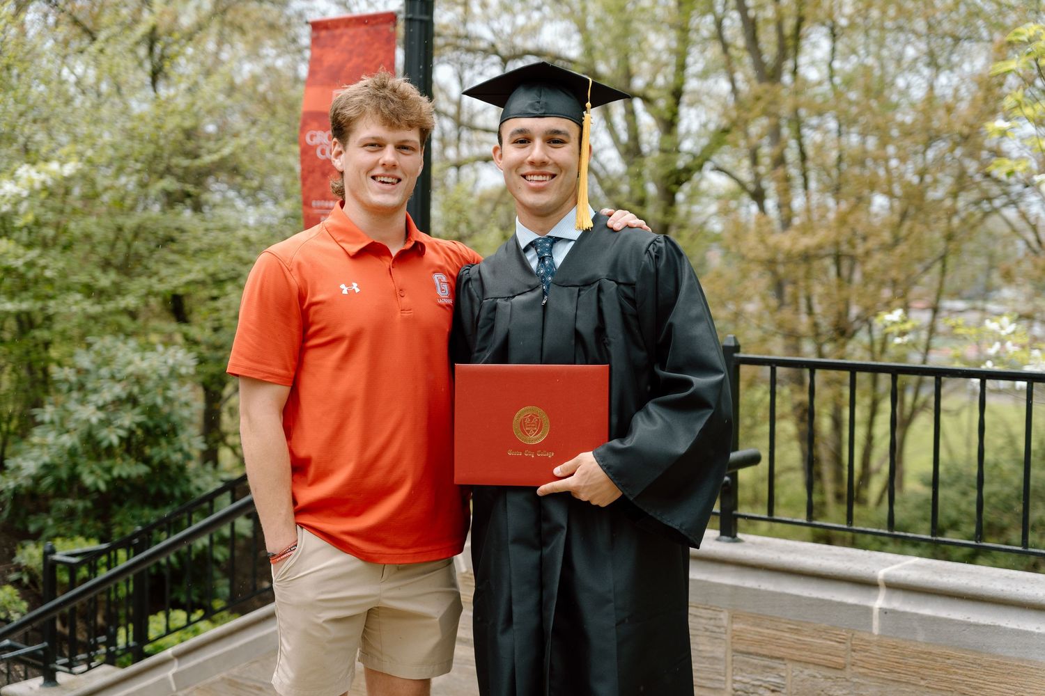 Graduate in cap and gown poses with family member holding orange diploma cover.