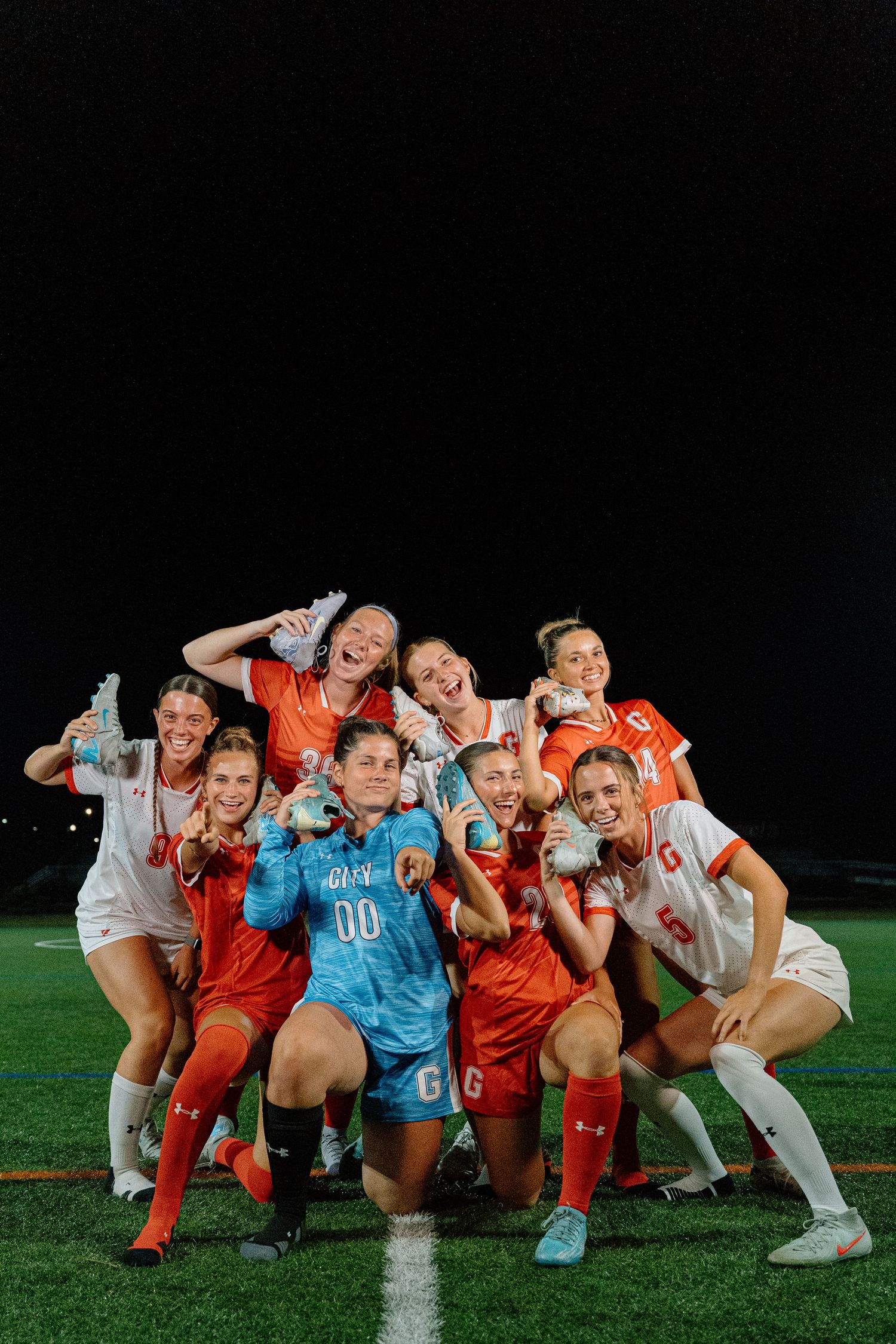 Happy soccer team in orange and white uniforms posing together on a sports field at night.