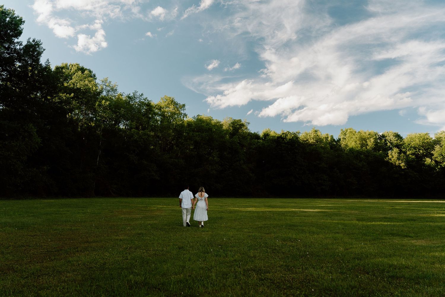 Couple in white attire walking hand in hand across a lush green field with trees and blue sky backdrop.