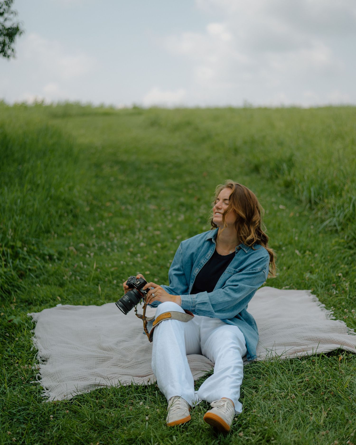 A photographer relaxes on a gray blanket in a lush green field while holding a camera.