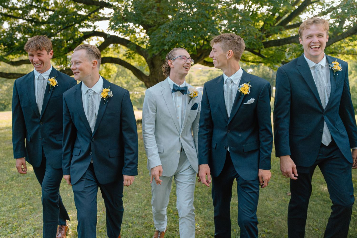 Groomsmen in navy and light grey suits smiling and walking together outdoors during wedding photos.