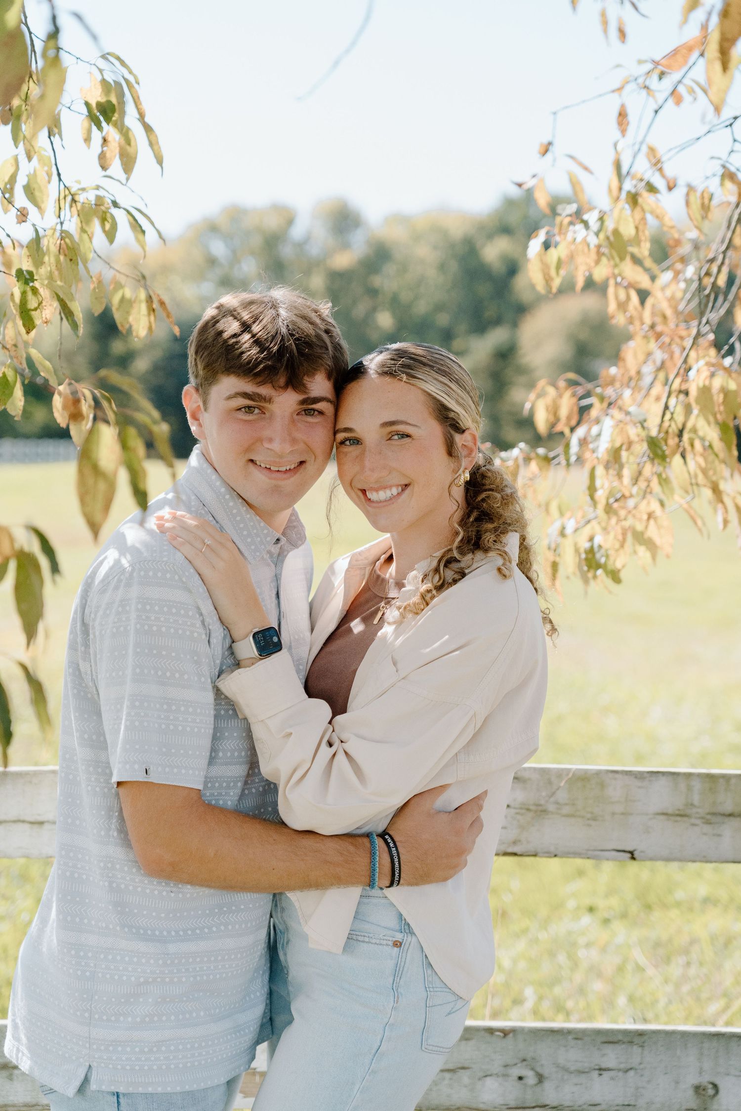 Two people embrace and smile while standing outdoors near a wooden fence with autumn trees in the background.