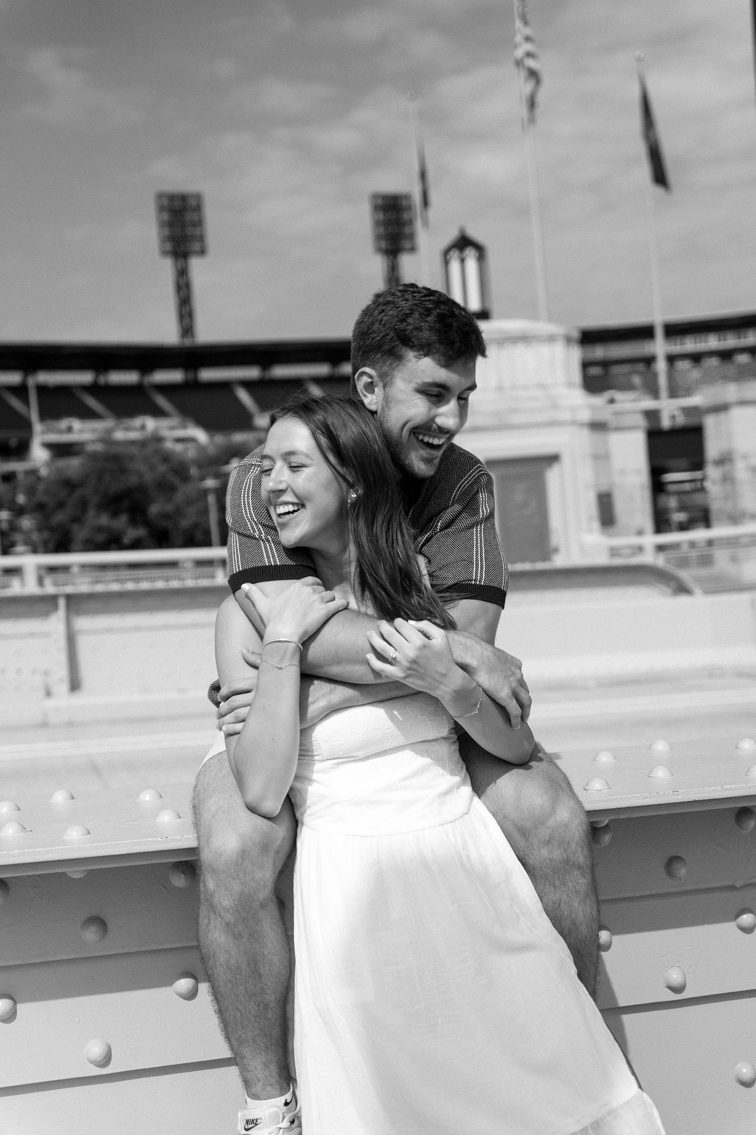 Black and white photo of a laughing couple embracing outdoors near a stadium on a sunny day.
