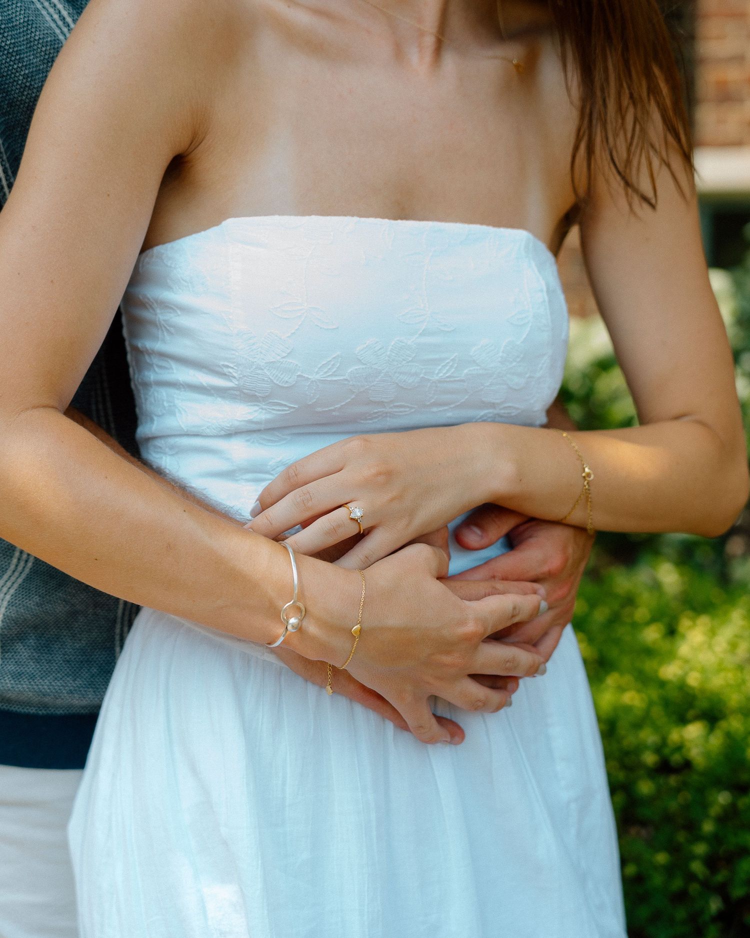 A couple embraces outdoors, showing hands with delicate jewelry against a white strapless dress.