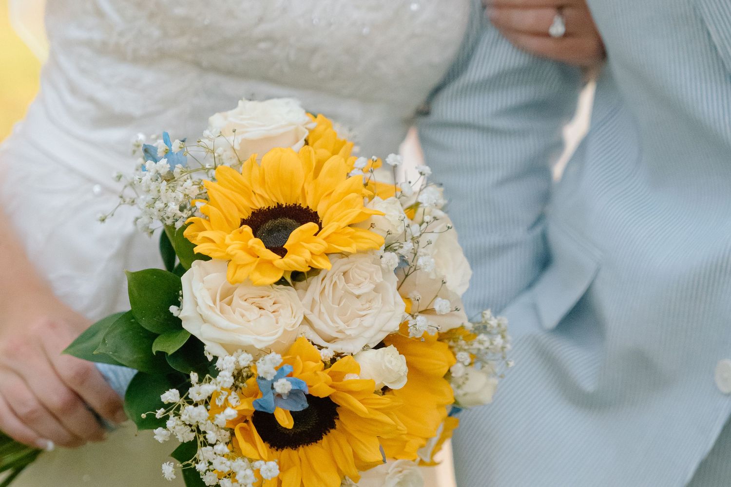 A wedding bouquet featuring bright yellow sunflowers and white roses with baby's breath accents against formal attire.