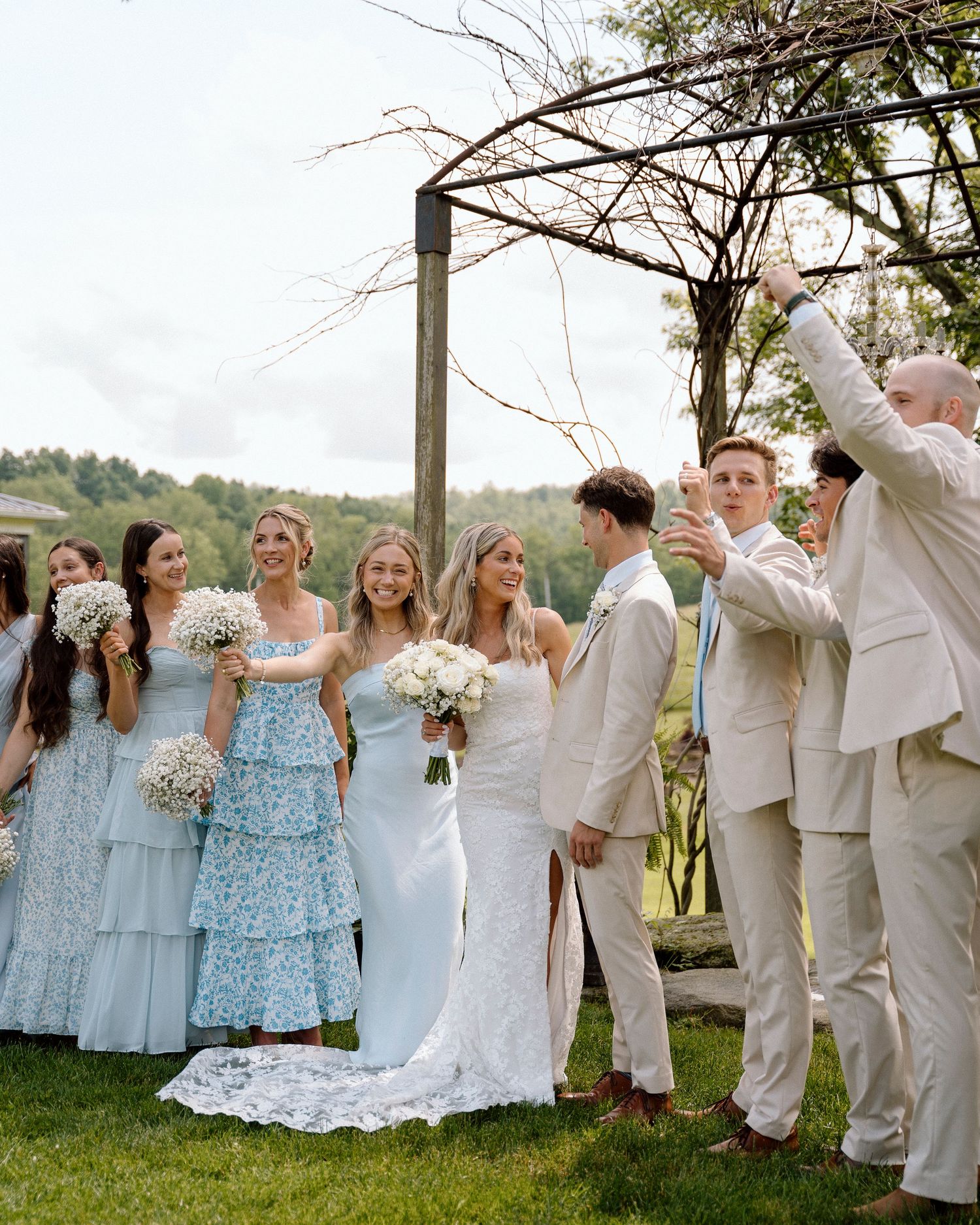 Wedding party in light blue dresses and beige suits pose together outdoors under wooden arch.