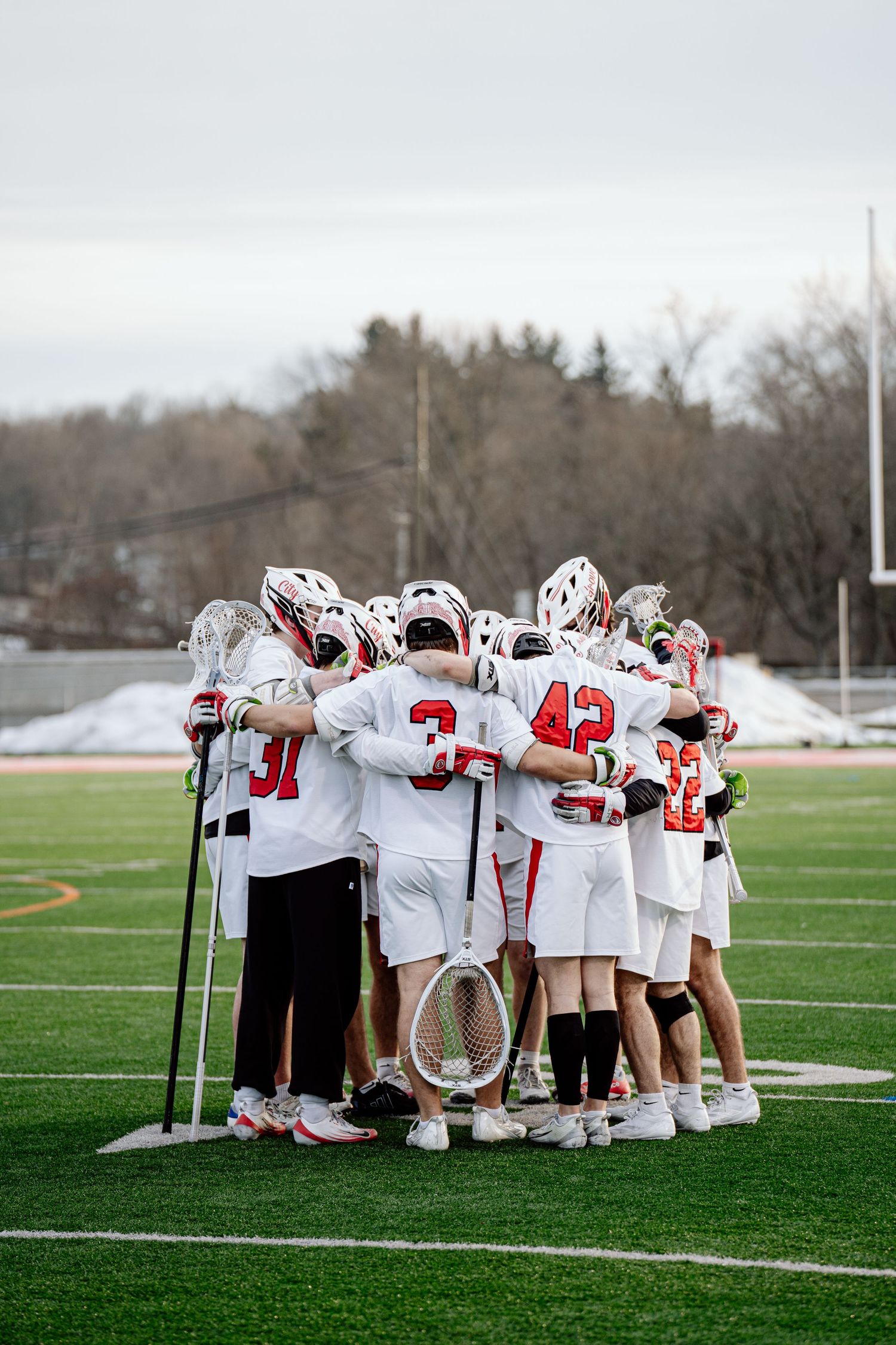Lacrosse team in white and red uniforms huddles together on a green turf field during a cold weather game.